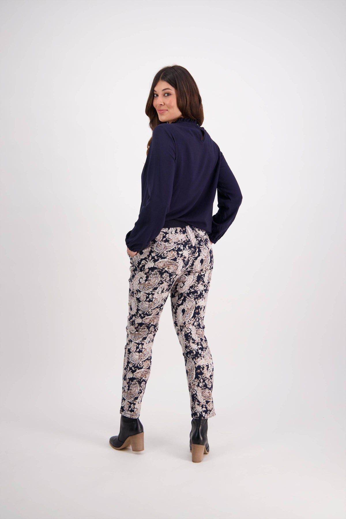A woman stands facing away, looking over her shoulder. She wears a navy long-sleeve top, Vassalli Ankle Grazer Pant - Antique, and heeled ankle boots, posing against a plain light background.
