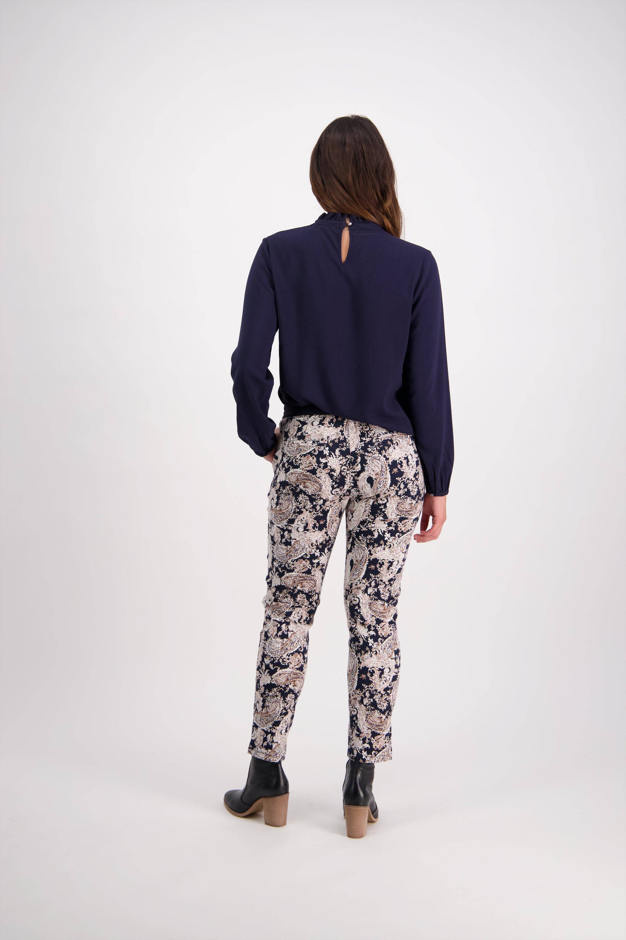 A woman stands facing away, wearing a long-sleeved navy blouse, Vassalli Ankle Grazer Pant - Antique with a floral pattern and stretch fabric, plus black ankle boots with tan heels against a plain white background.