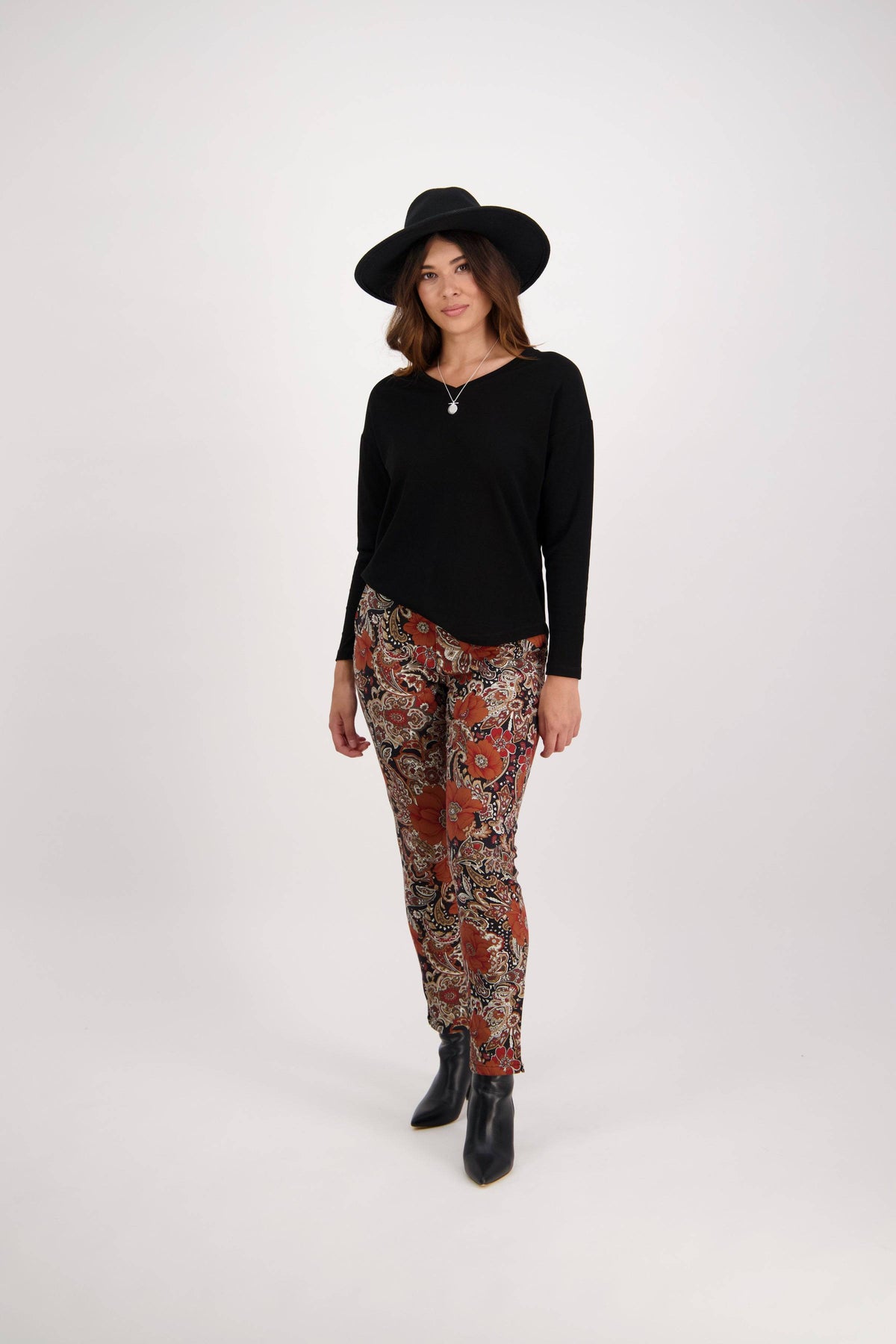 A woman stands against a plain white background, wearing a black wide-brimmed hat, black long-sleeve top, Vassalli Ankle Grazer Pant - Bohemian in red and beige, and black ankle boots.