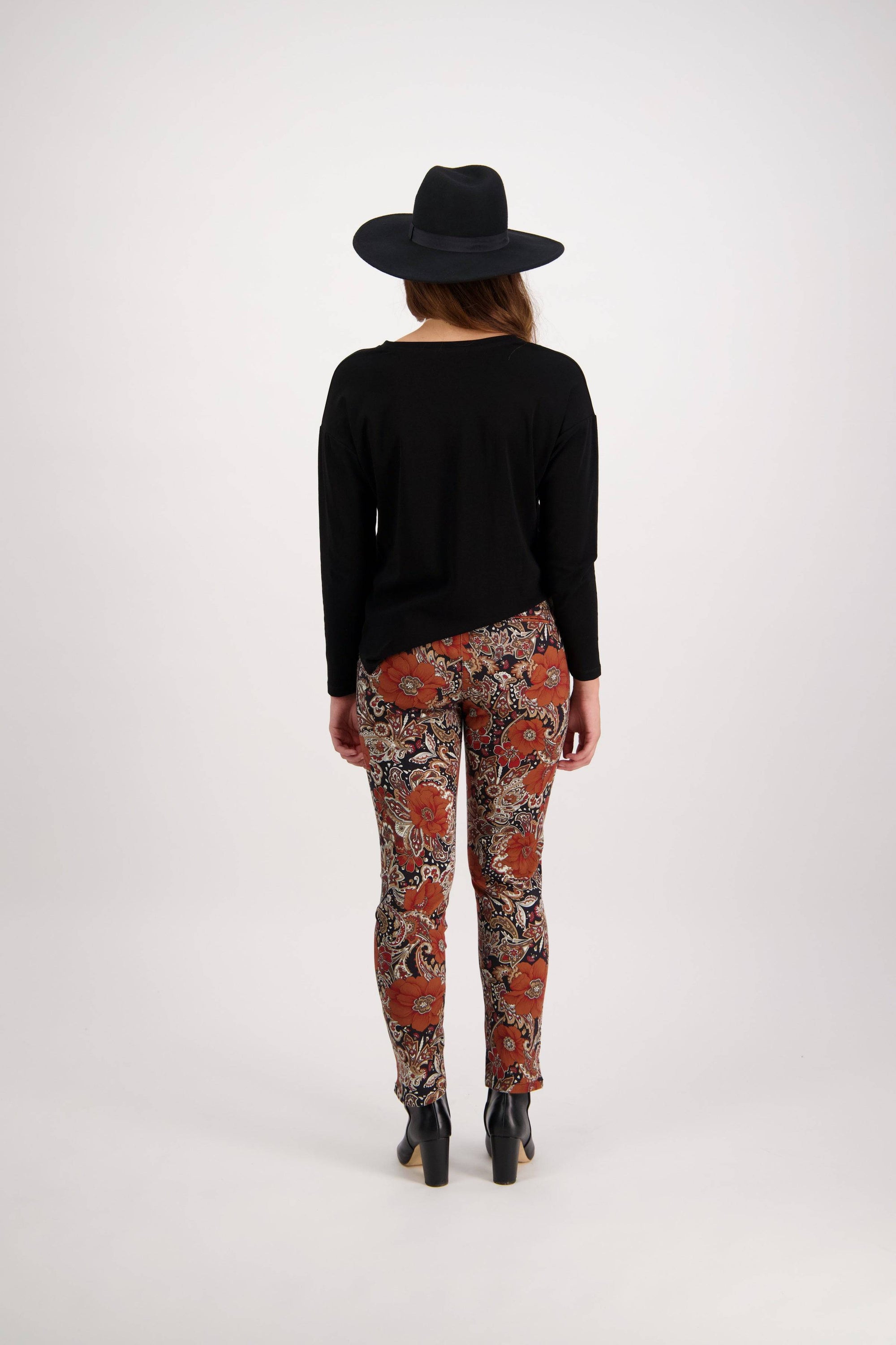 A person stands with their back to the camera, wearing a black wide-brim hat, black long-sleeve top, Vassalli Ankle Grazer Pant - Bohemian featuring a colorful floral print, and black heeled ankle boots against a plain white background.