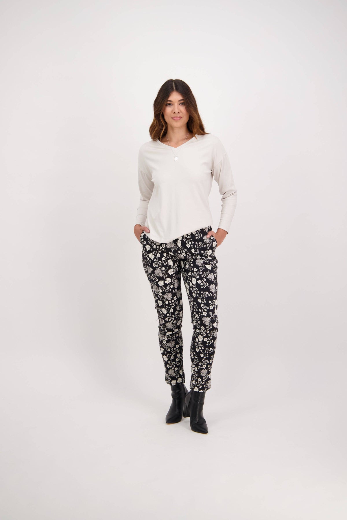A woman stands against a white background, wearing a long-sleeve white top, Vassalli Ankle Grazer Pant - Gypsy with a black and white floral print, and black ankle boots, hands in her pockets and smiling slightly.