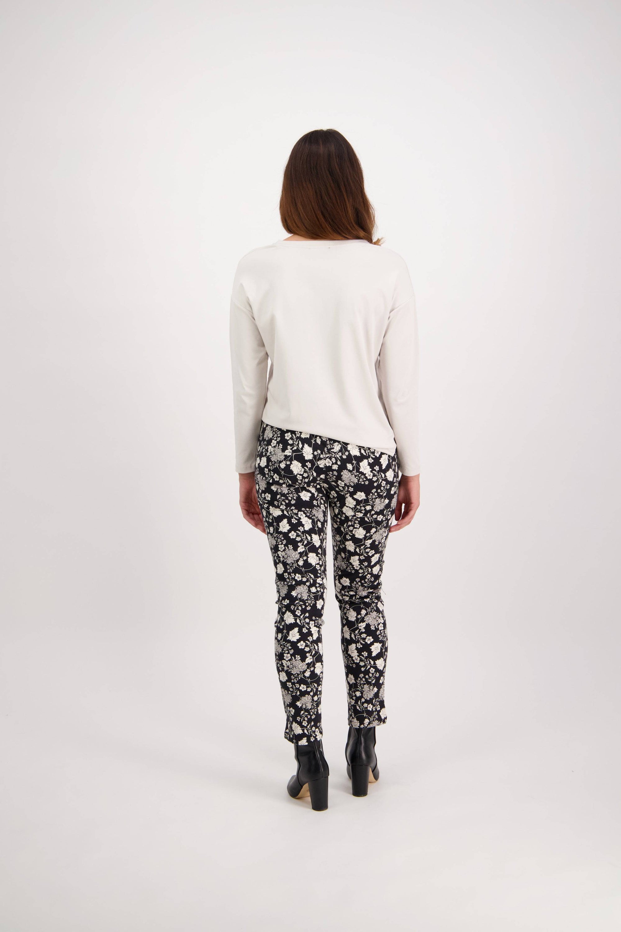 A woman faces away from the camera, wearing a white long-sleeve top, Vassalli Ankle Grazer Pant - Gypsy, and black heeled ankle boots against a plain white background.