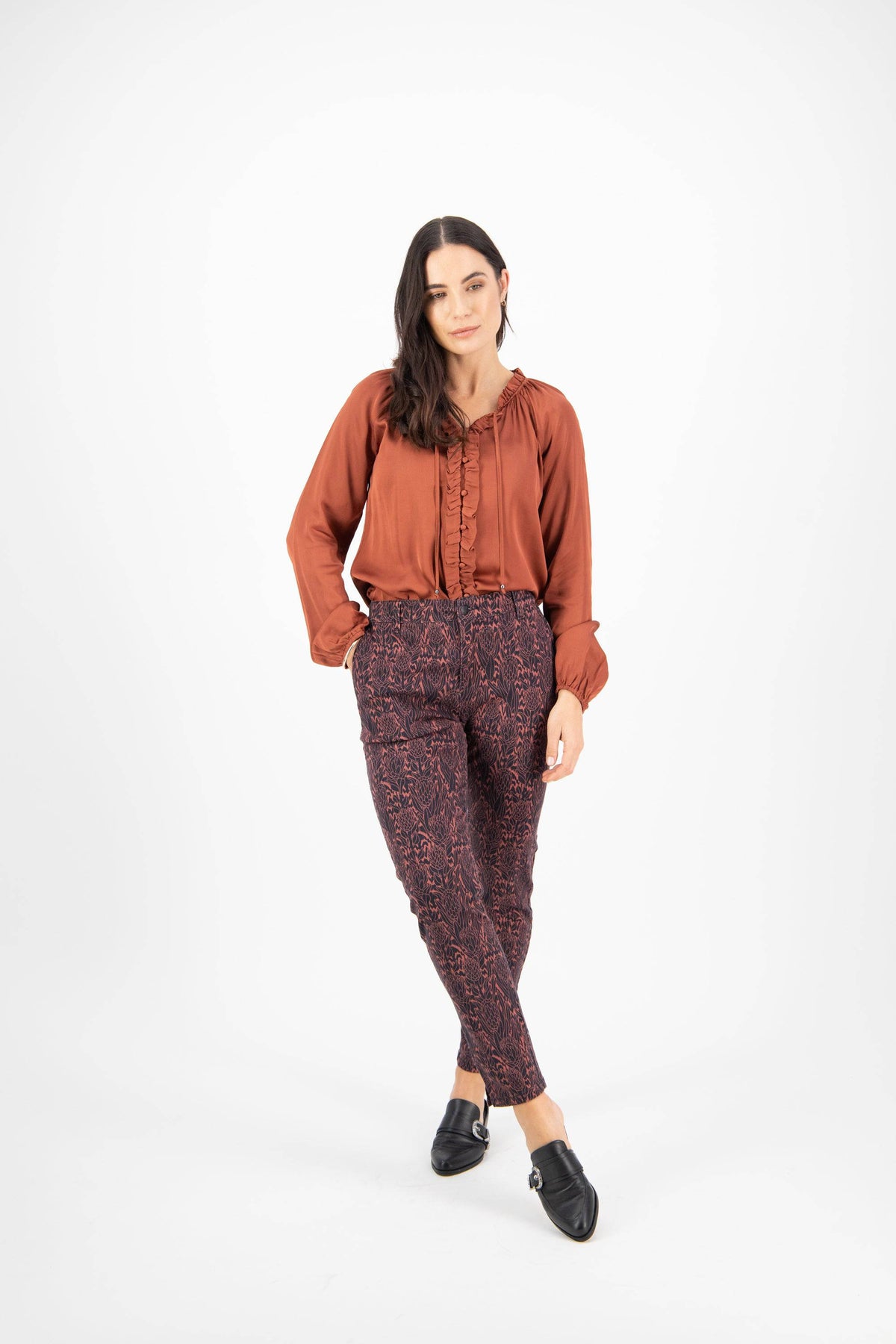A woman with long dark hair poses confidently against a white background, wearing Vassalli's Ankle Grazer Pant - Protea, a rust long-sleeve ruffle blouse, and black slip-on shoes.