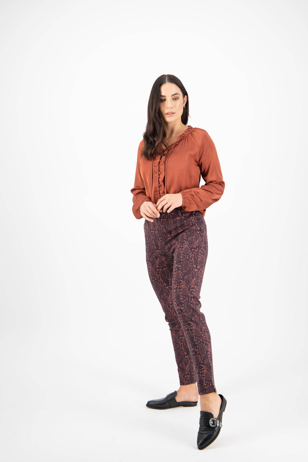 A woman with dark hair stands against a white background, wearing a rust ruffle blouse, Vassalli Ankle Grazer Pant - Protea in dark tones, and black loafers, looking slightly to the side.