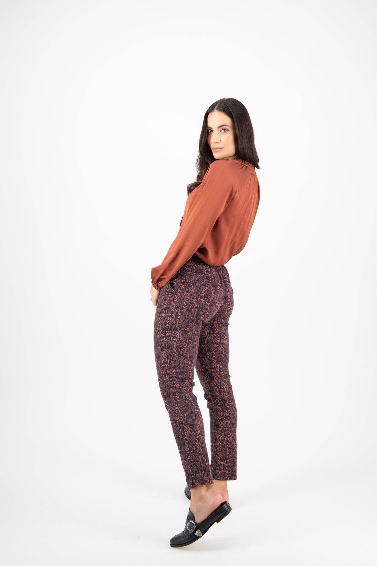 Woman with long dark hair wearing a rust blouse, Vassalli Ankle Grazer Pant - Protea, and black loafers, standing and looking over her shoulder against a plain white background.
