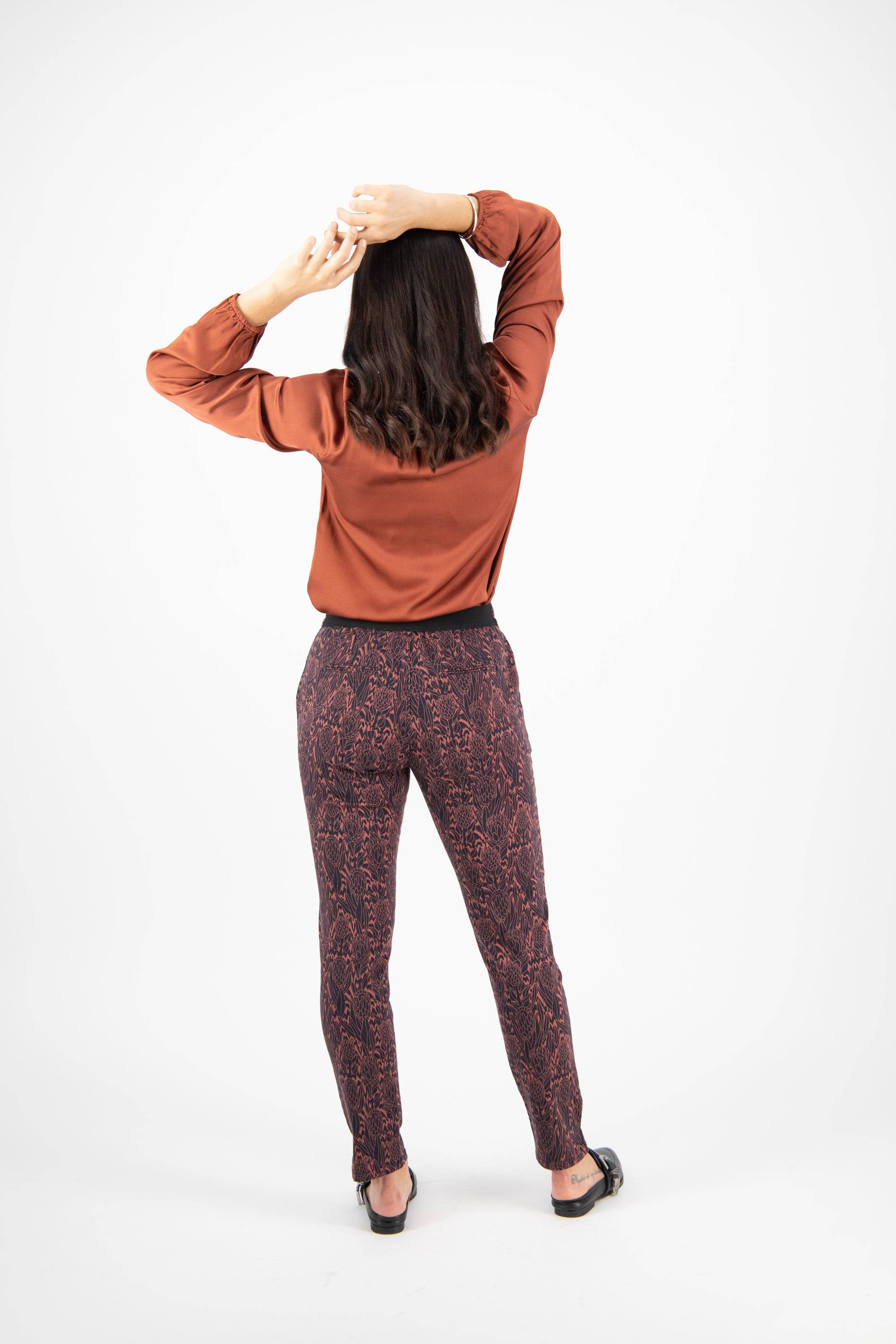 A woman faces away with arms raised, wearing a rust long-sleeve top, Vassalli Ankle Grazer Pant - Protea, and black sandals against a plain white background.