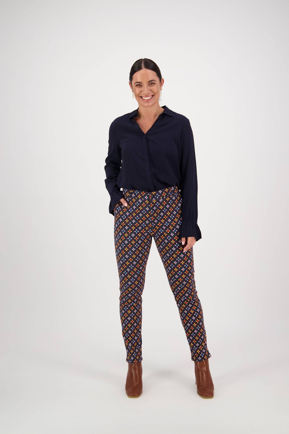 A woman smiles against a white background, wearing a dark long-sleeve blouse and Vassalli Ankle Grazer Pant - Stellar, featuring a colorful geometric print. Made from stretch fabric, these pants pair perfectly with brown ankle boots. One hand is in her pocket.