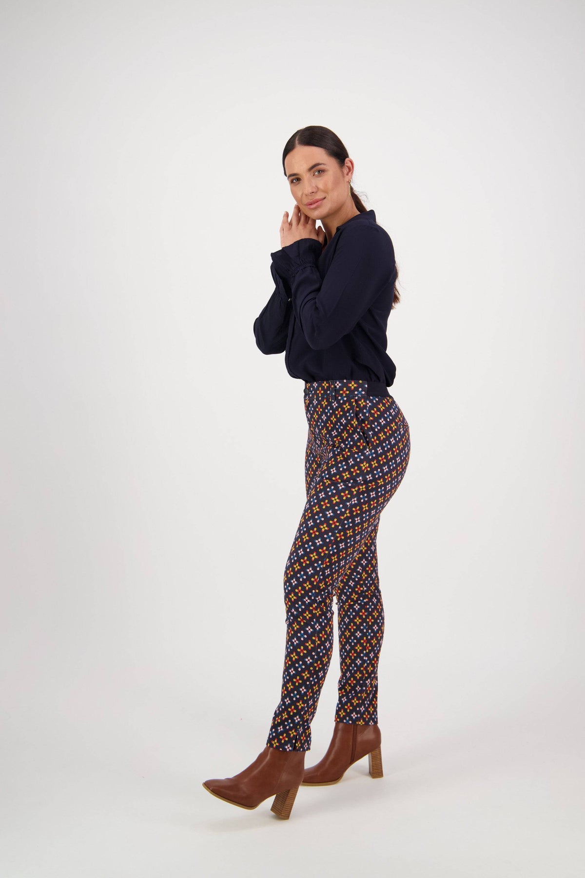A woman with long dark hair, in a navy blouse, Vassalli Ankle Grazer Pant - Stellar, and brown heeled boots poses sideways against a plain white background.