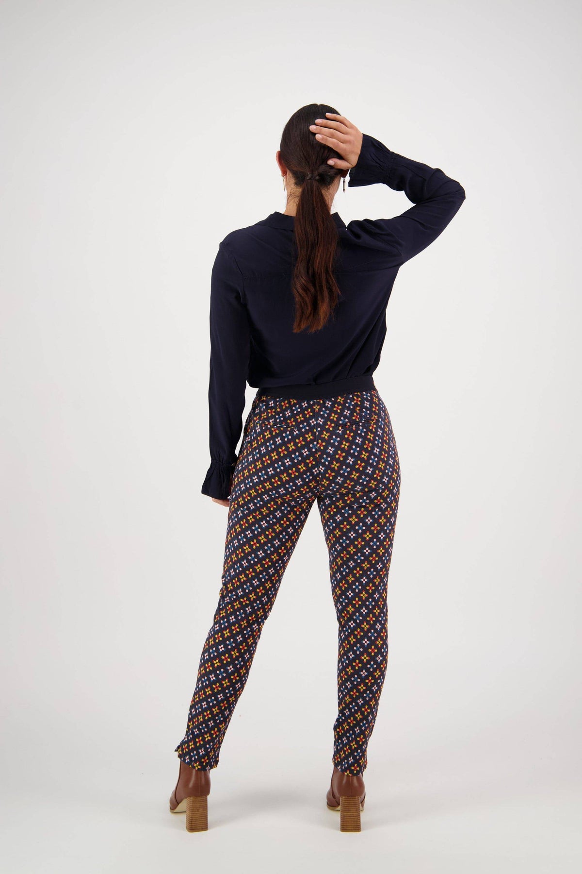 A woman faces away, holding her ponytail. She wears a dark long-sleeve top, Vassalli Ankle Grazer Pant - Stellar in an orange and blue print stretch fabric, with brown heeled shoes against a plain white background.