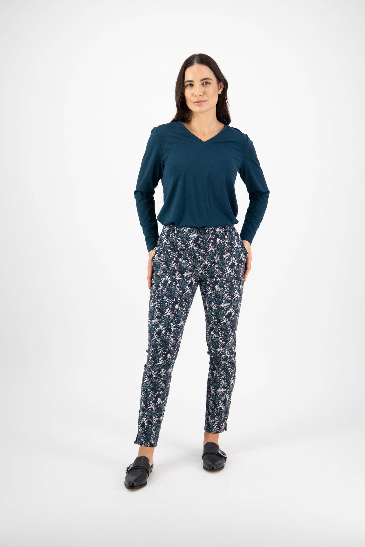 A woman stands facing forward with her hands in her pockets, wearing a teal long-sleeve top, black slip-on shoes, and Vassalli Ankle Grazer Pant - Aspen against a plain white background.
