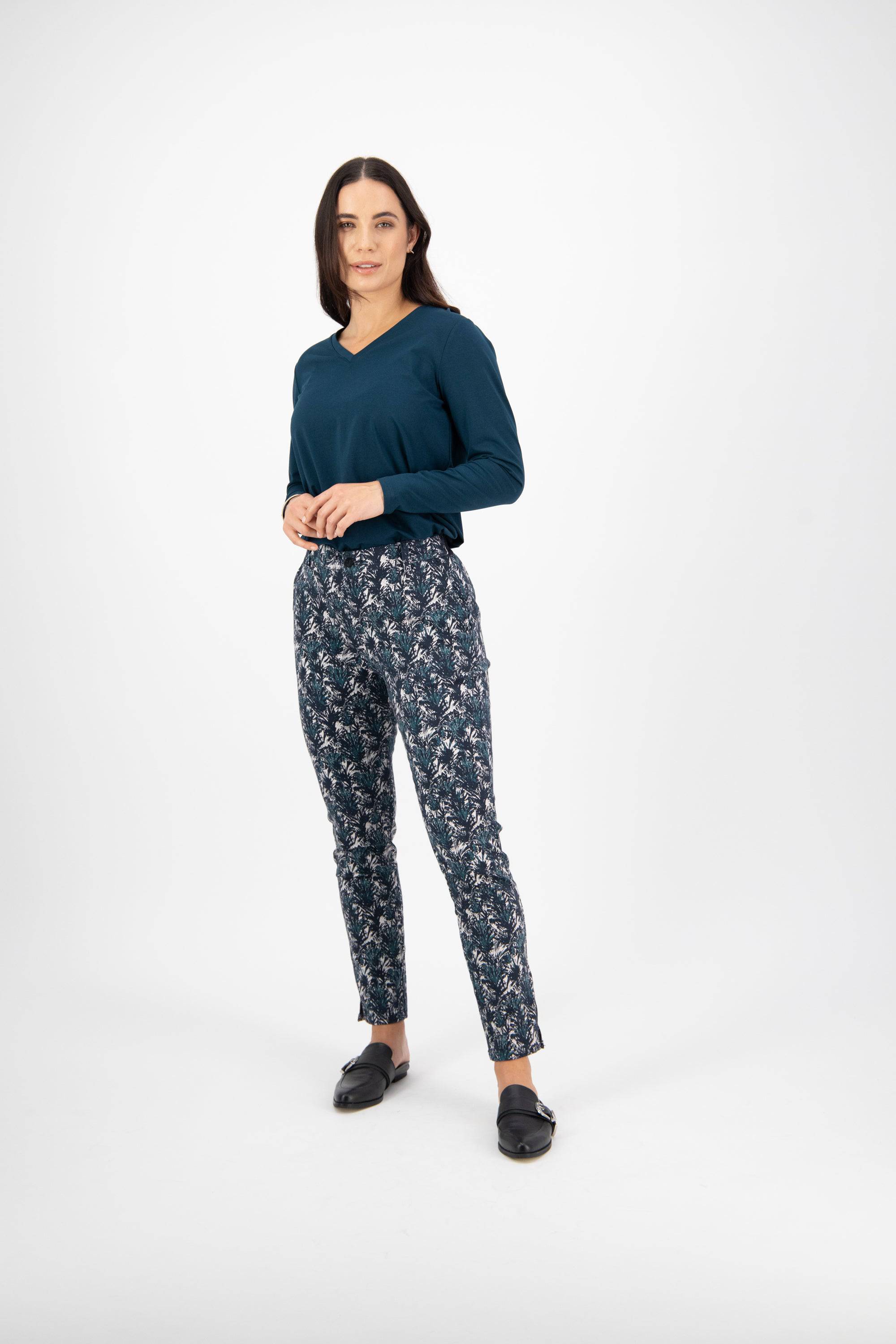 A woman with long brown hair stands against a white background wearing a teal top, black slip-on shoes, and Vassalli Ankle Grazer Pant - Aspen in blue and white, gazing at the camera with a neutral expression.