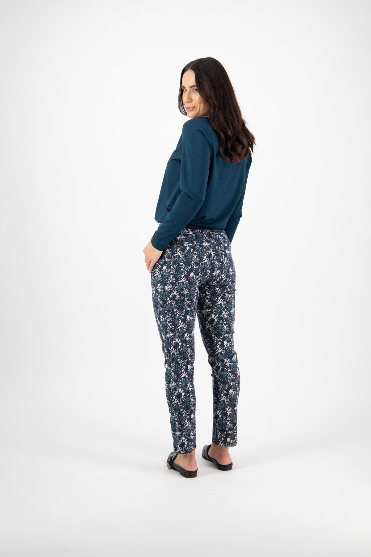 A woman with long dark hair stands facing away, looking over her shoulder. She wears a teal long-sleeve top, Vassalli Ankle Grazer Pant - Aspen, and black sandals against a plain white background.