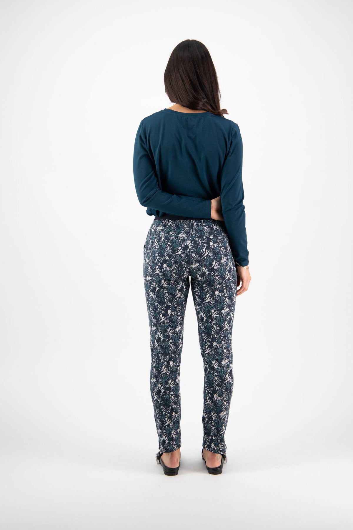 A woman with long dark hair faces away, wearing a dark blue long-sleeve top and Vassalli Ankle Grazer Pant - Aspen. She stands on a white background with her arms relaxed at her sides.