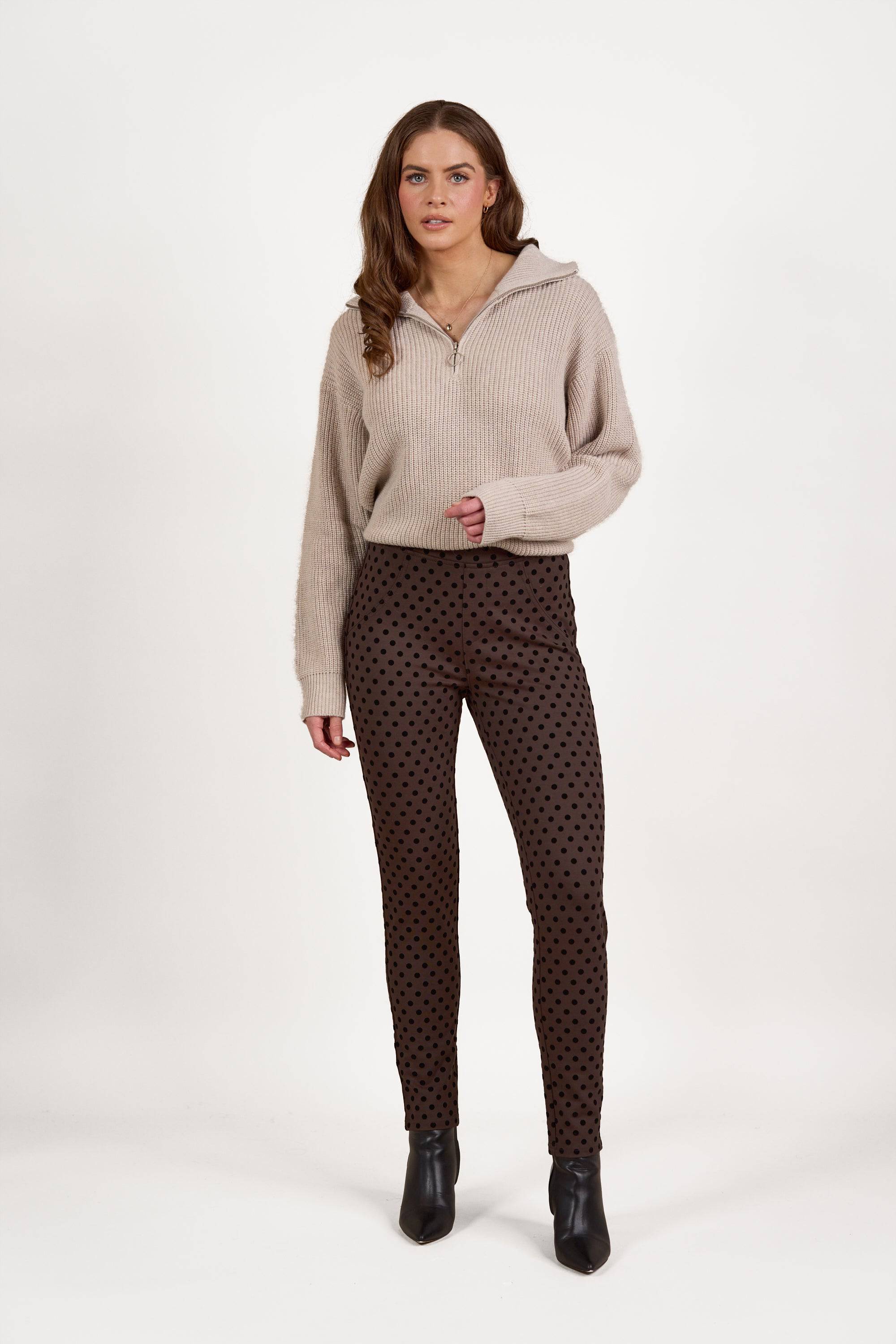 A woman with long brown hair and a neutral expression stands against a white background wearing Vassalli's View Pant in Chocolate with a skinny leg ankle grazer fit, paired with a beige zip-up sweater and black ankle boots.
