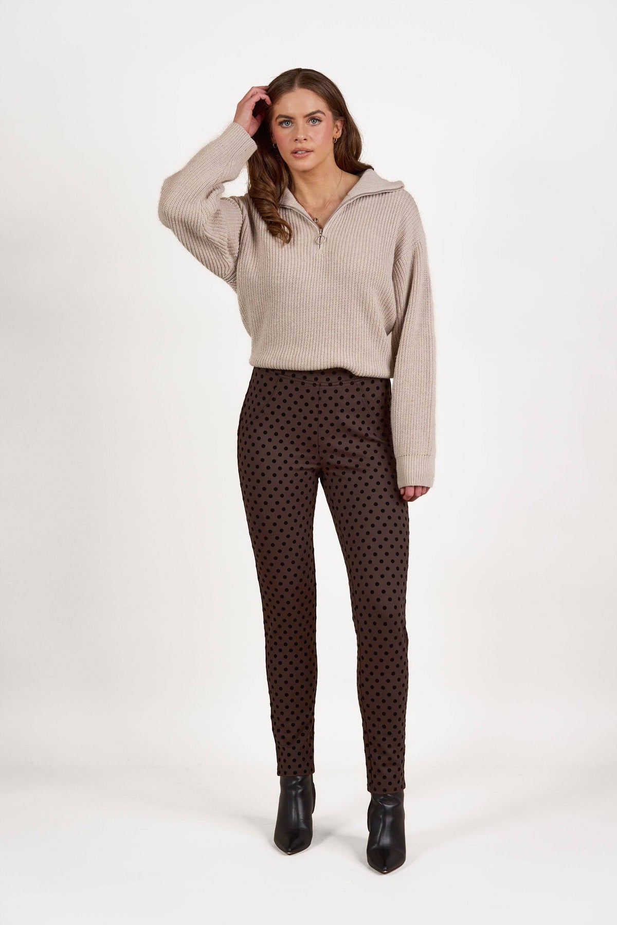 A woman stands against a white background wearing the Vassalli View Pant - Chocolate and a beige zip-up sweater, with black ankle boots. She has her left hand touching her hair.