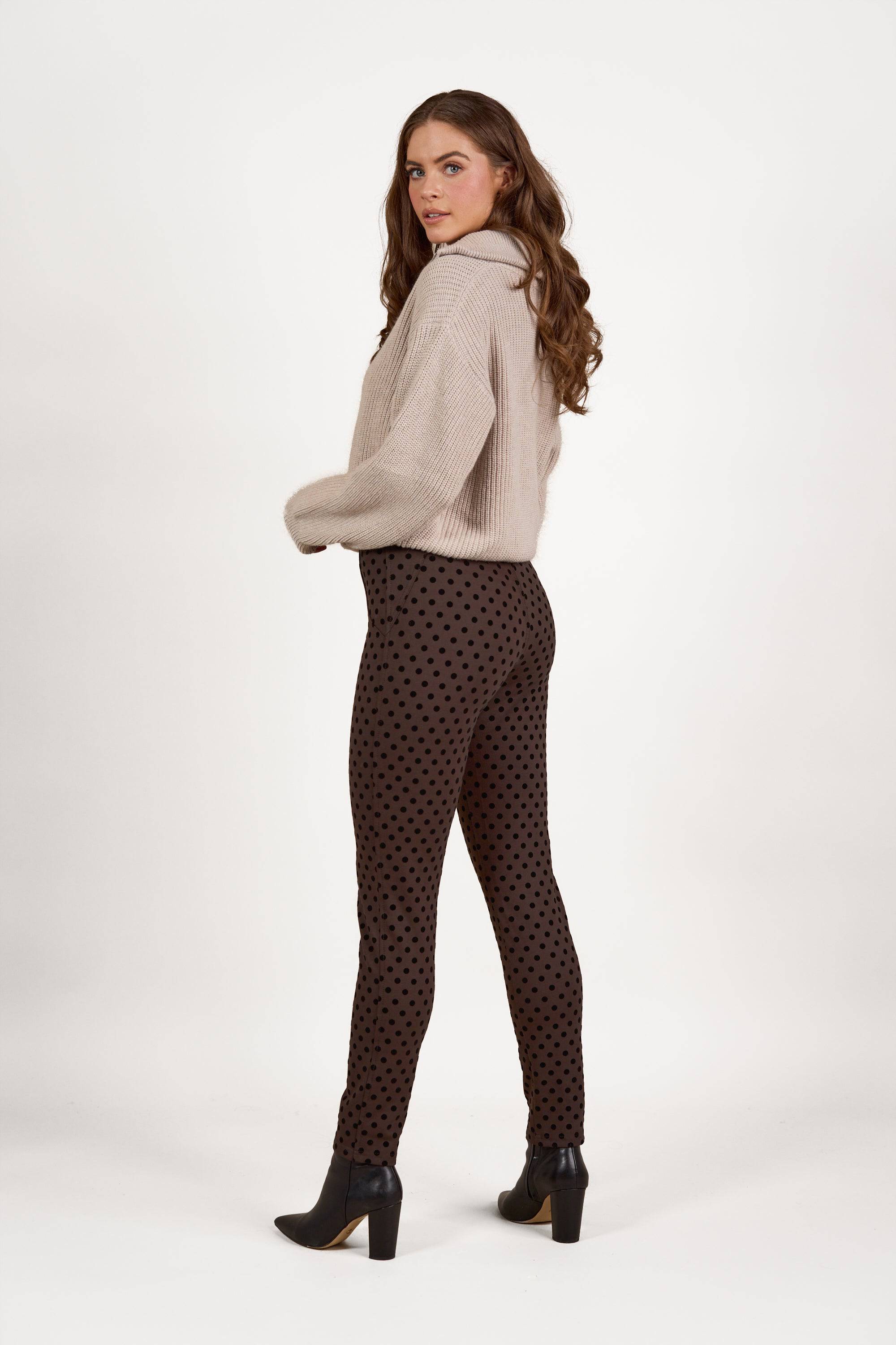 A woman with long brown hair models the Vassalli View Pant in Chocolate, styled with a beige sweater and black heeled ankle boots. She stands sideways against a white background, looking over her shoulder.