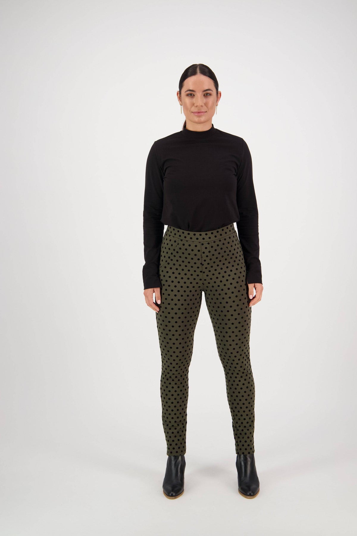 A woman stands facing forward wearing a black long-sleeve top, Vassalli View Pant - Khaki with black polka dots, and black ankle boots against a plain white background.