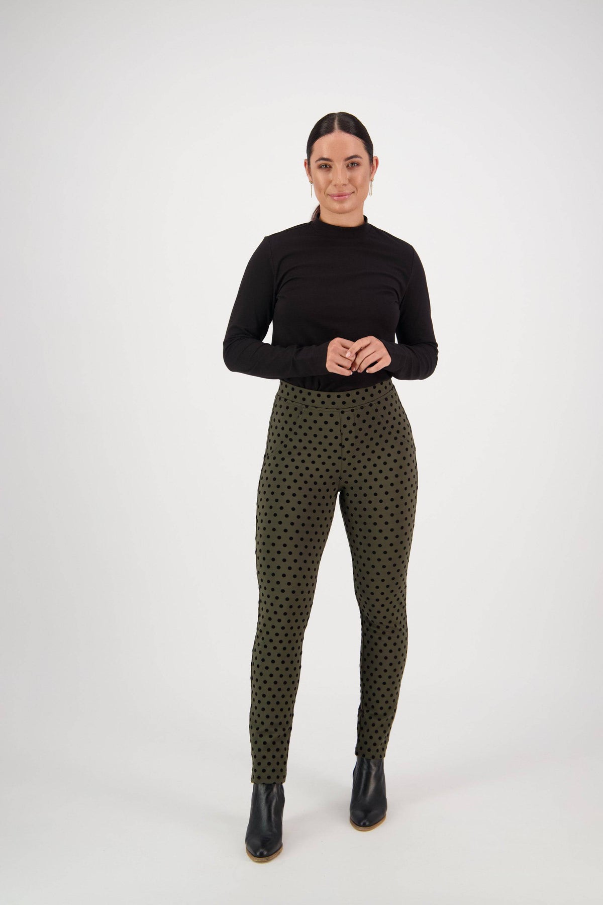 A person stands facing forward against a plain white background, wearing a black long-sleeve top, Vassalli View Pant in khaki with black polka dots, and black ankle boots.