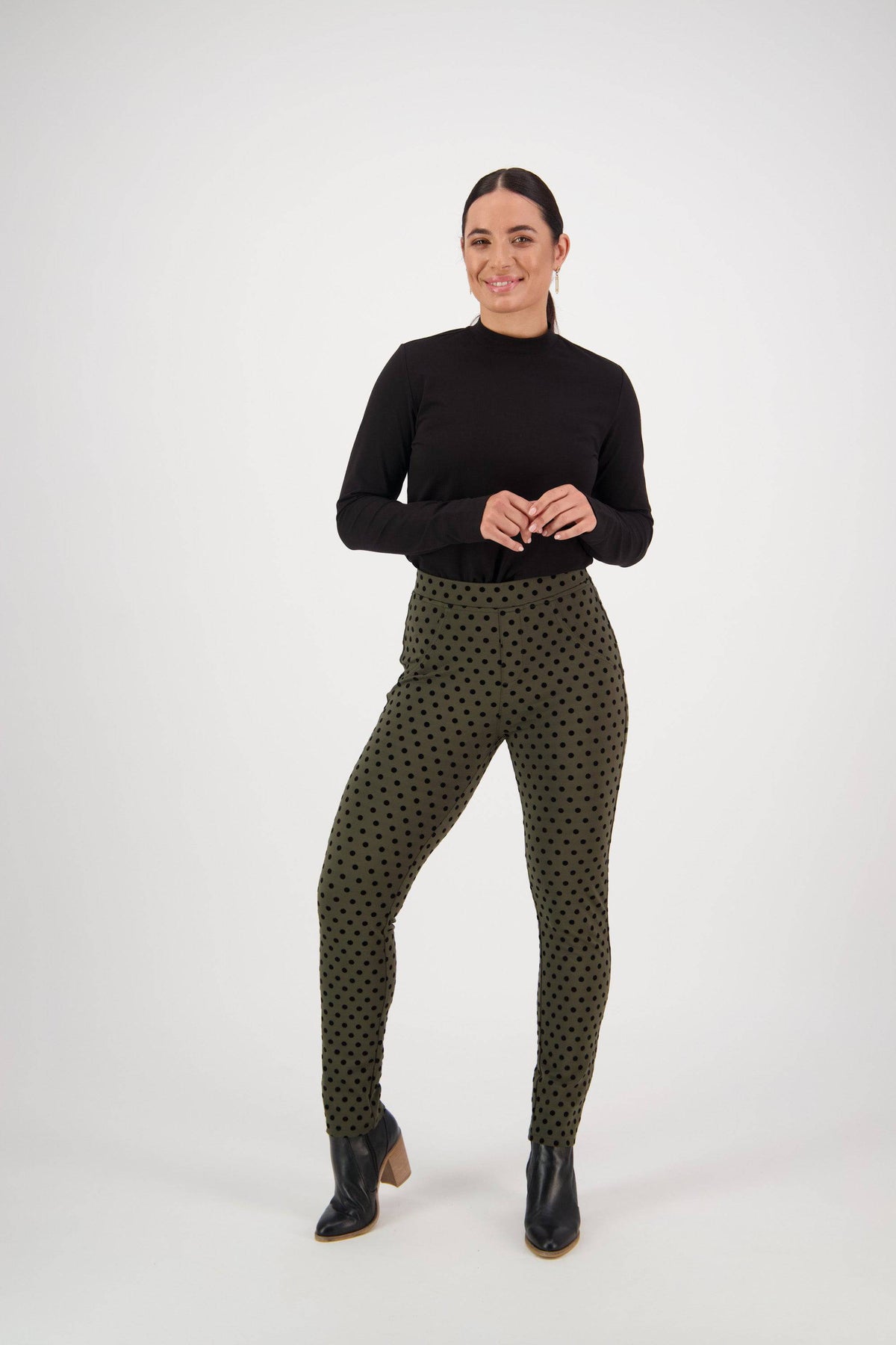 A woman stands against a white background, smiling in her Vassalli View Pant - Khaki, paired with a black long-sleeve top and black ankle boots. She clasps her hands in front of her.