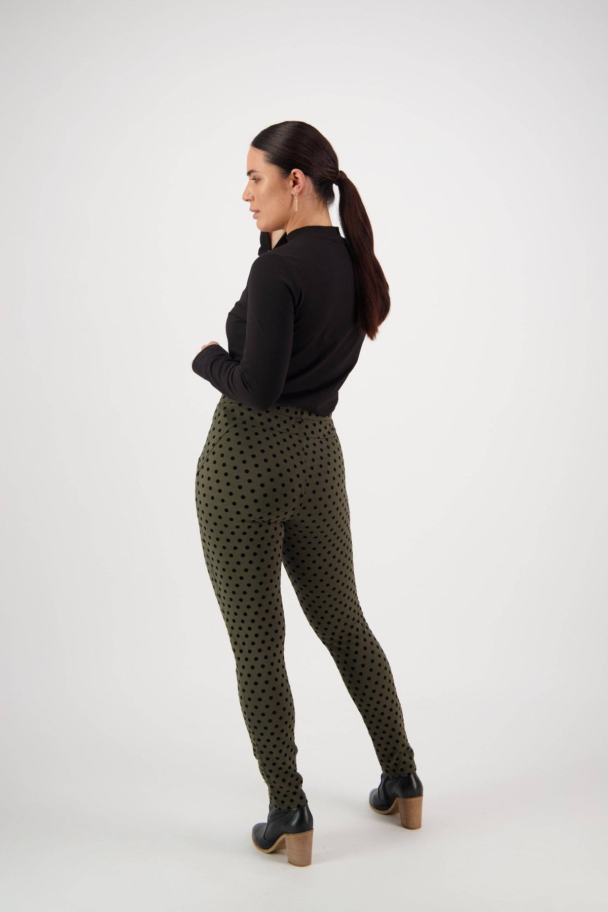 A woman with a ponytail stands in a three-quarter pose, wearing a black long-sleeve top, Vassalli View Pant - Khaki with black polka dots, and black shoes with wooden heels against a plain white background.