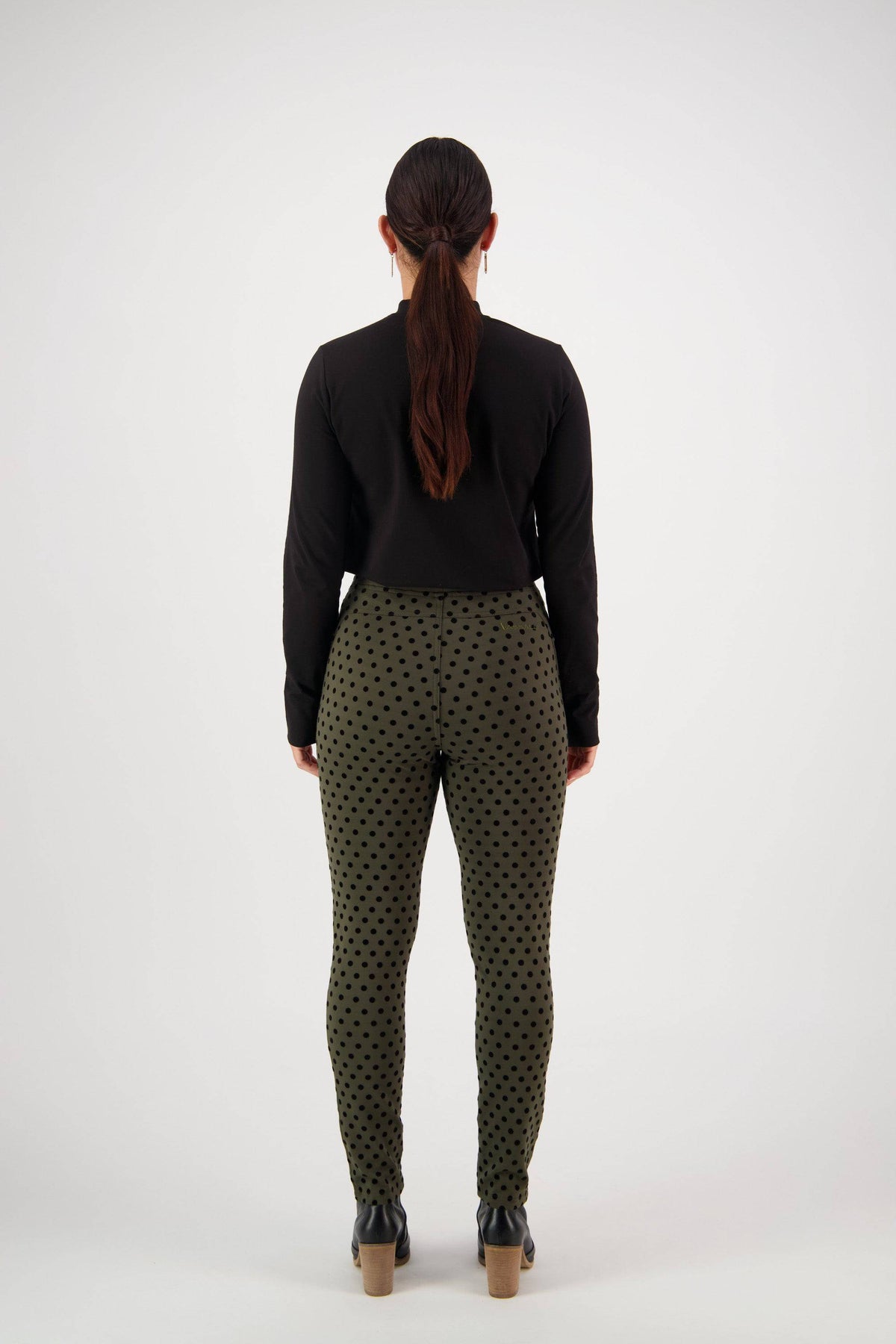 A person with long brown hair in a ponytail stands facing away, wearing a black long-sleeve top, Vassalli View Pant - Khaki with black polka dots, and black shoes against a plain white background.