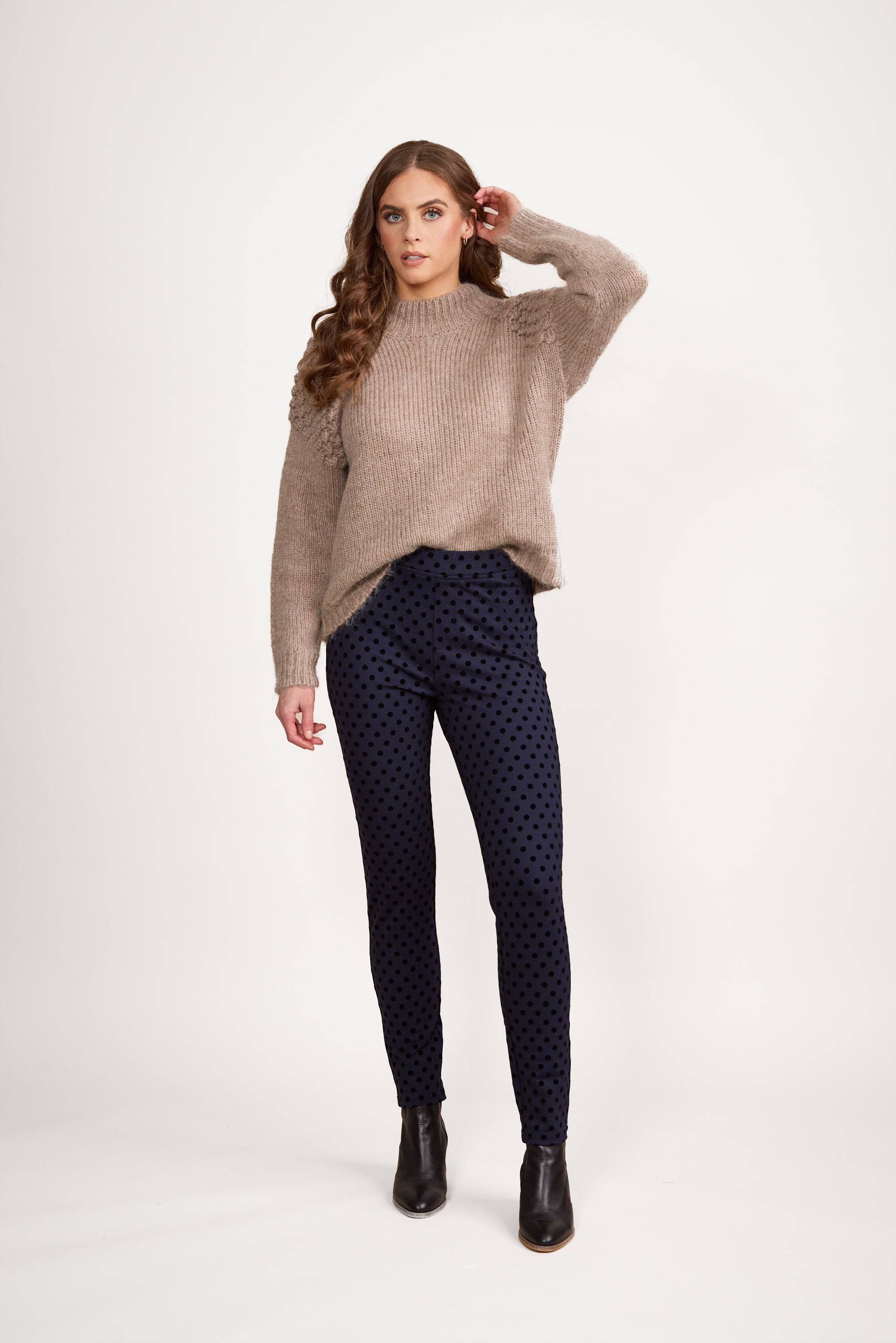 A woman with long brown hair wears a loose beige sweater, Vassalli View Pant in Navy, and black ankle boots, standing against a plain white background. She has one hand near her ear and looks at the camera.