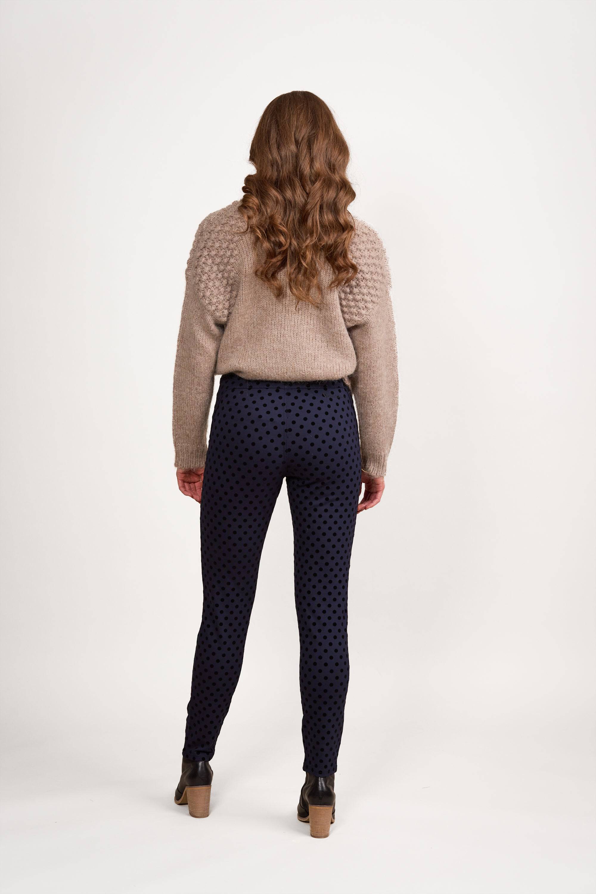 A person with long, wavy brown hair faces away from the camera, wearing a beige textured sweater and Vassalli View Pant - Navy with brown heeled ankle boots, against a white background.