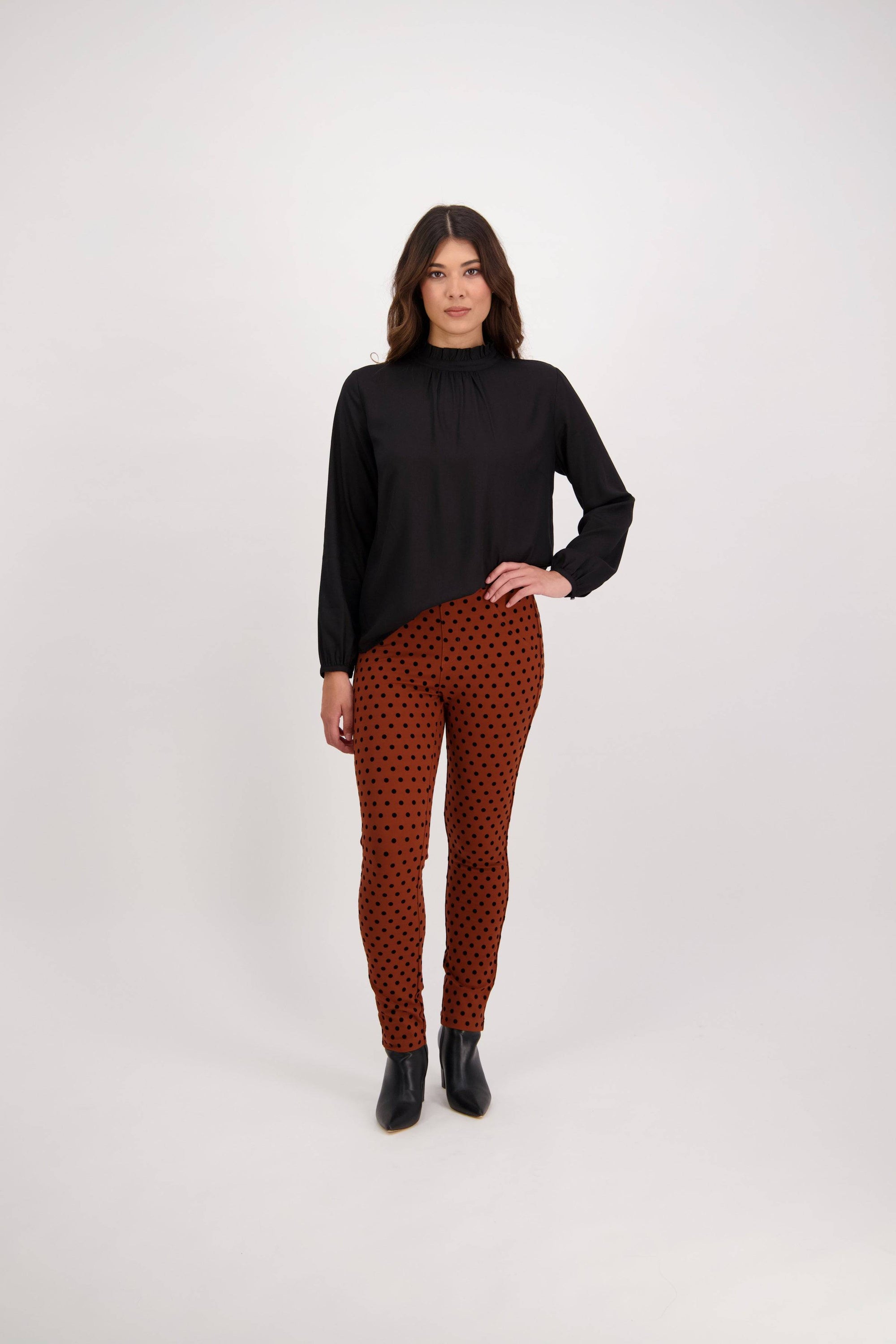 A woman with shoulder-length dark hair stands against a white background, wearing a long-sleeve black top and Vassalli’s View Pant - Rustic, styled with black ankle boots. She faces the camera, her rust-colored pants featuring subtle black dots.