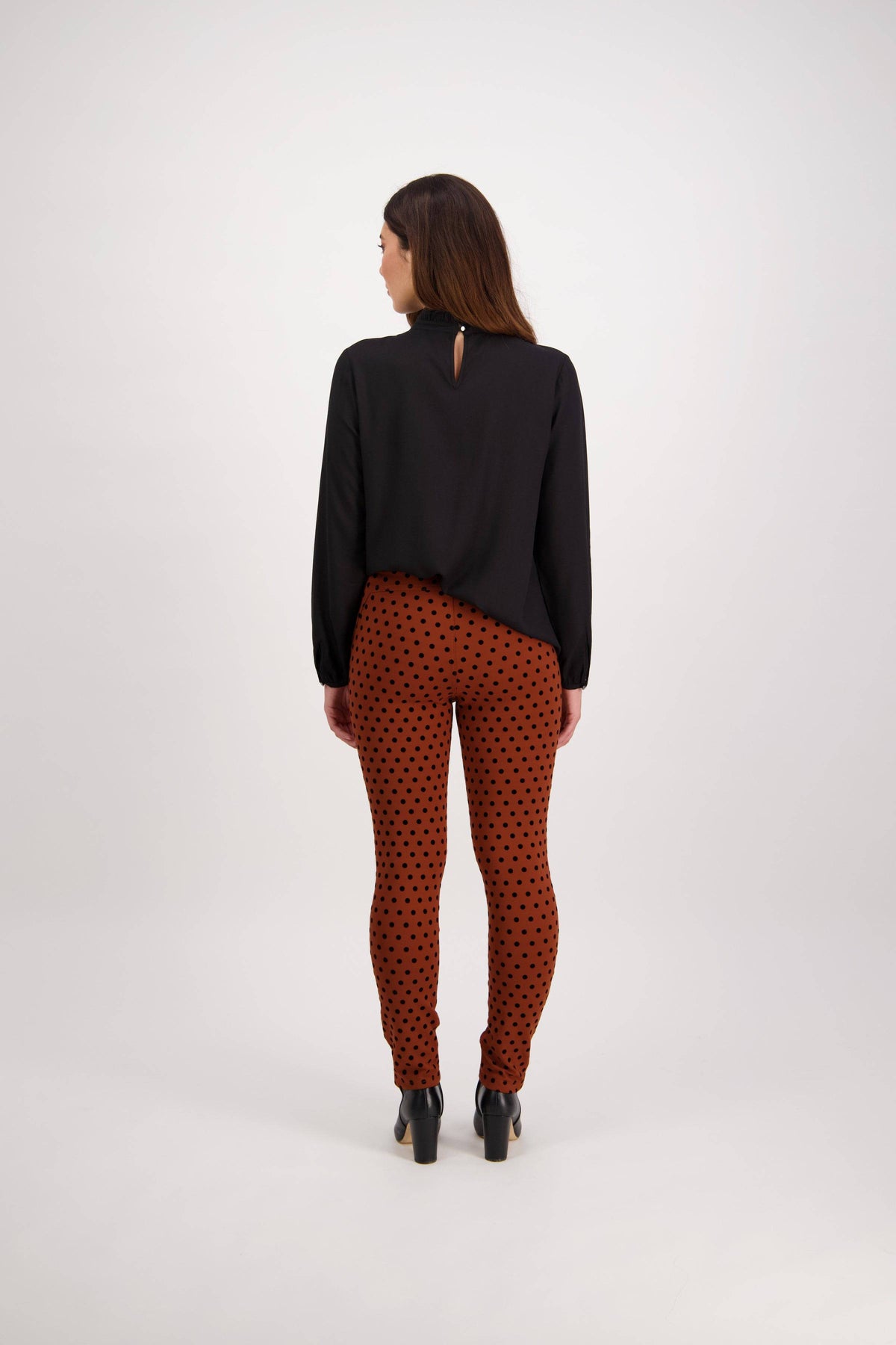A woman with long brown hair faces away from the camera, wearing the Vassalli View Pant - Rustic and a black long-sleeve top, standing against a plain white background.