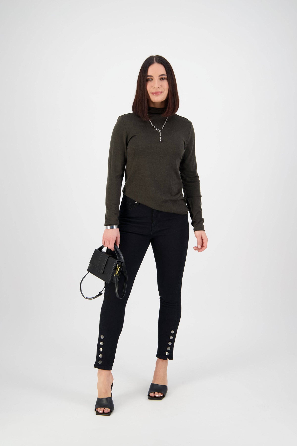 A woman with straight, shoulder-length dark hair stands against a plain white background, wearing a dark green sweater, Vassalli Ankle Grazer Jean - Black Denim, black heeled sandals, and holding a small black handbag.
