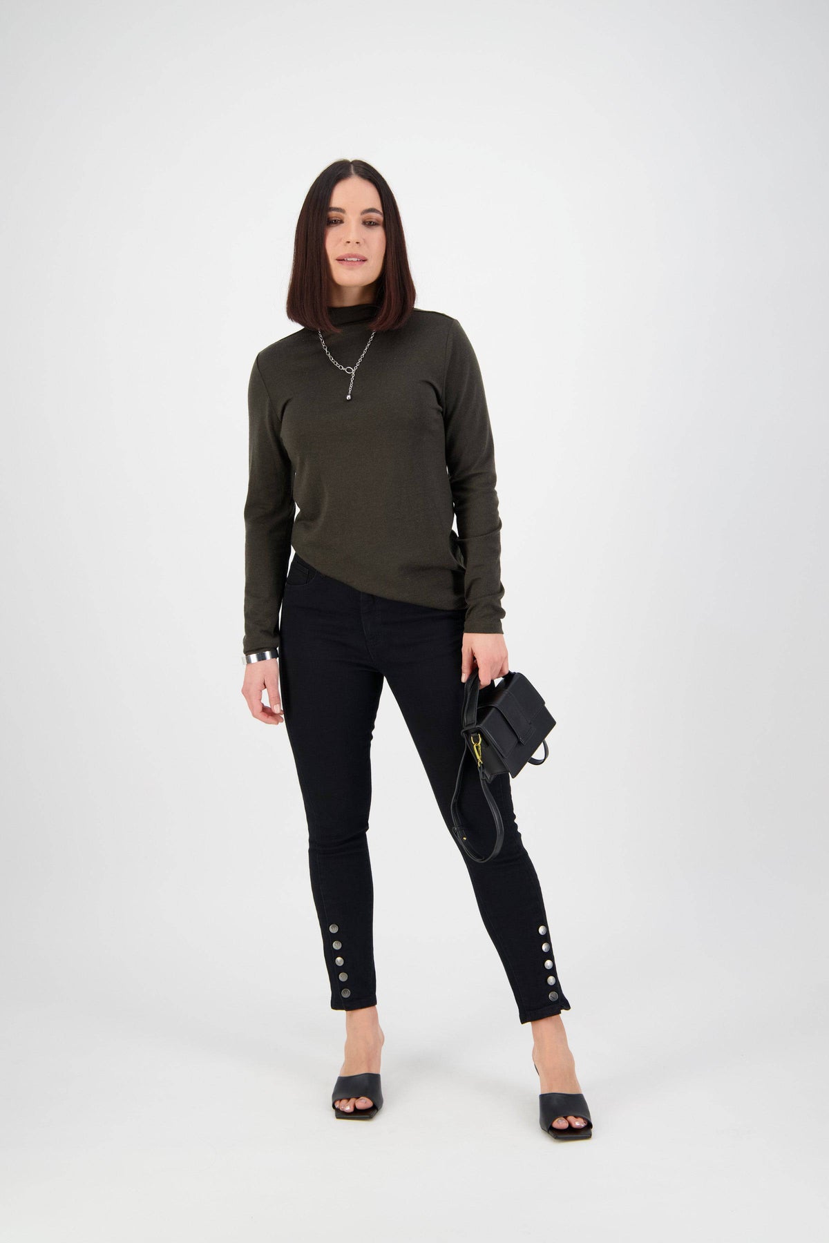 A woman with straight dark hair stands against a white background, wearing a dark green long-sleeve top, Vassalli Ankle Grazer Jean - Black Denim, black heeled sandals, and holding a black handbag.