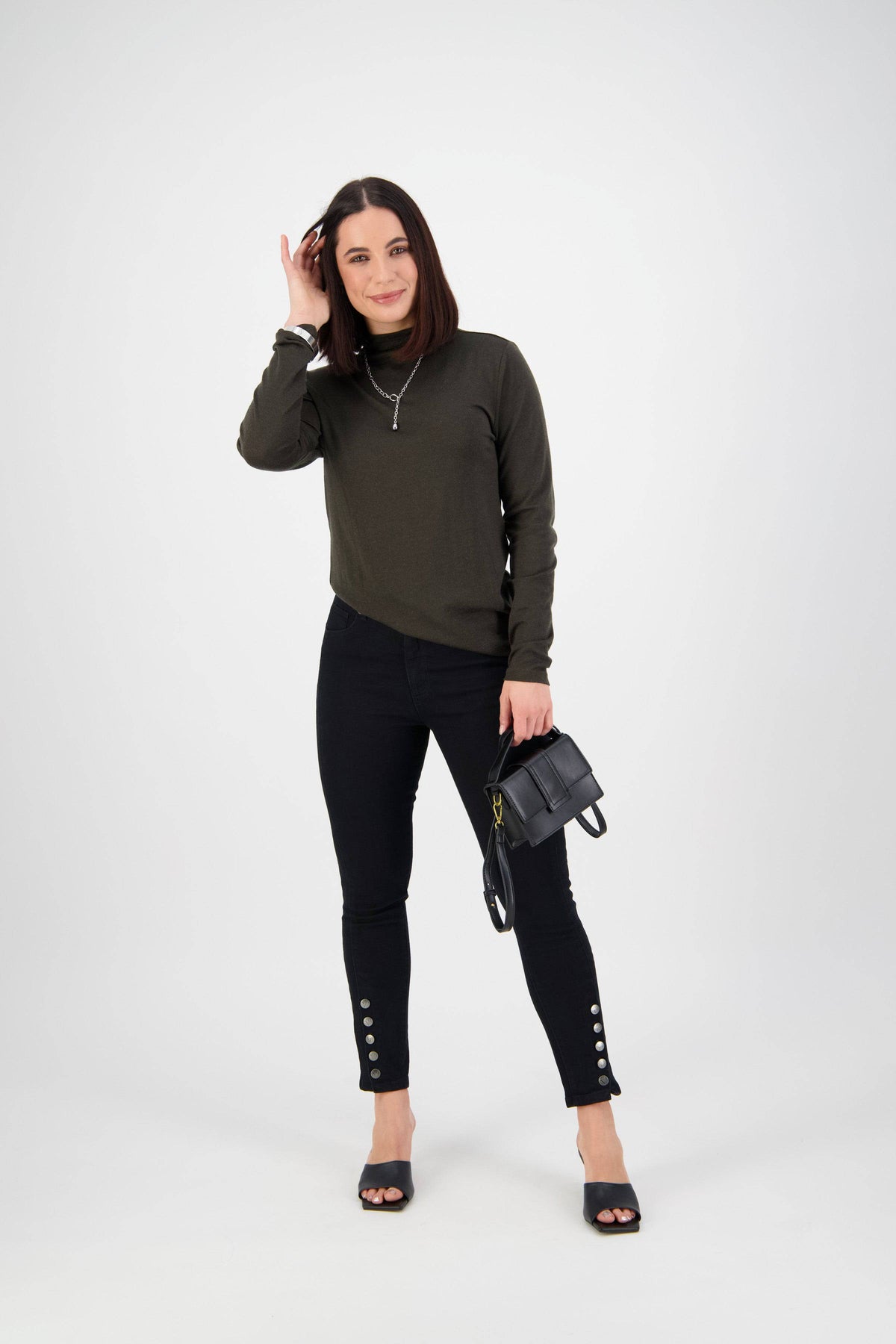 A woman with straight brown hair wears a dark green long-sleeve top, Vassalli Ankle Grazer Jean - Black Denim, and black open-toe shoes. She holds a small black handbag and smiles against a plain white background.