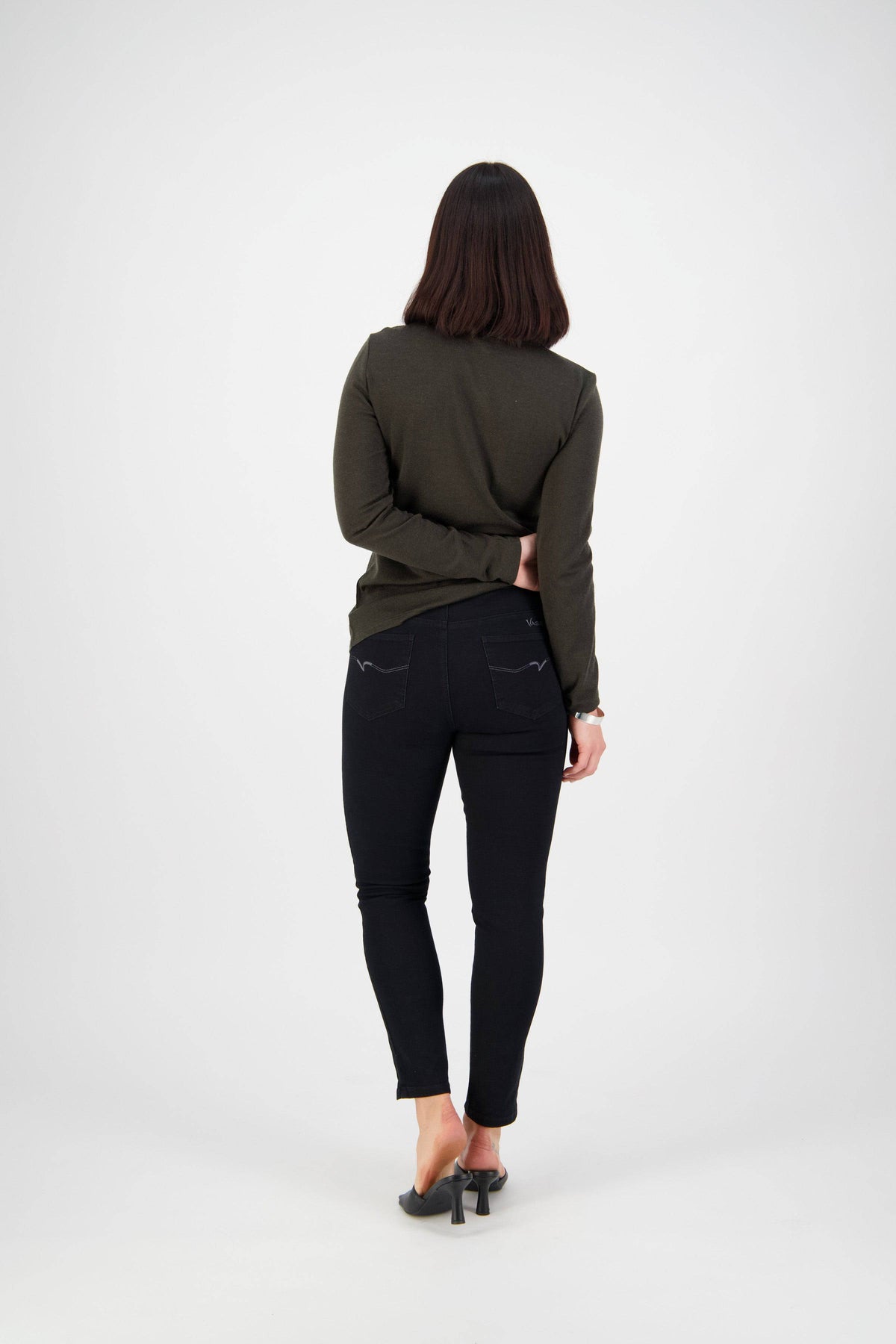 A woman with straight, shoulder-length dark hair faces away against a white background, wearing a dark green long-sleeve top, black heeled shoes, and Vassalli Ankle Grazer Jean in Black Denim with one arm bent behind her back.