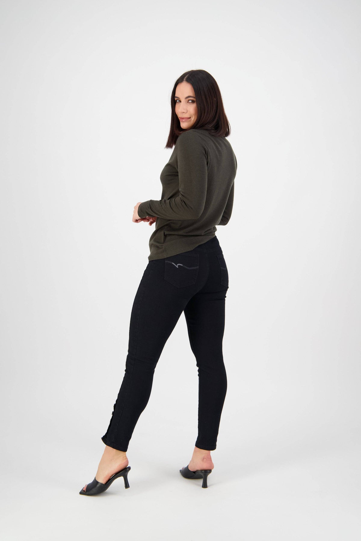 A woman with straight dark hair, in a dark green long-sleeve top, Vassalli Ankle Grazer Jean - Black Denim, and black heeled sandals stands facing away but looks back and smiles in a white studio.