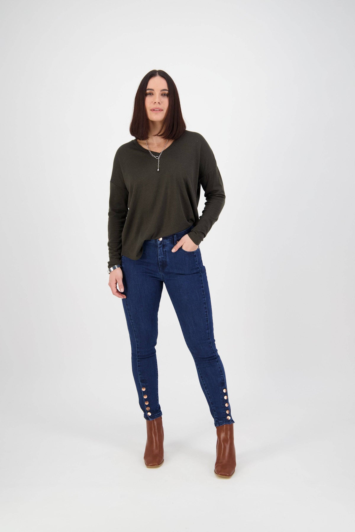 A woman with straight brown hair stands against a white background, wearing a dark green long-sleeve top, brown ankle boots, and Vassalli Ankle Grazer Jean in Ink Denim. She has one hand in her pocket.