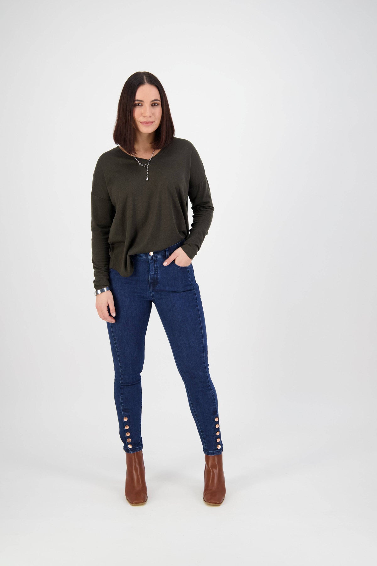 A woman with straight, shoulder-length brown hair stands against a white background, wearing a dark green long-sleeve top, Vassalli Ankle Grazer Jean - Ink Denim, and brown ankle boots.