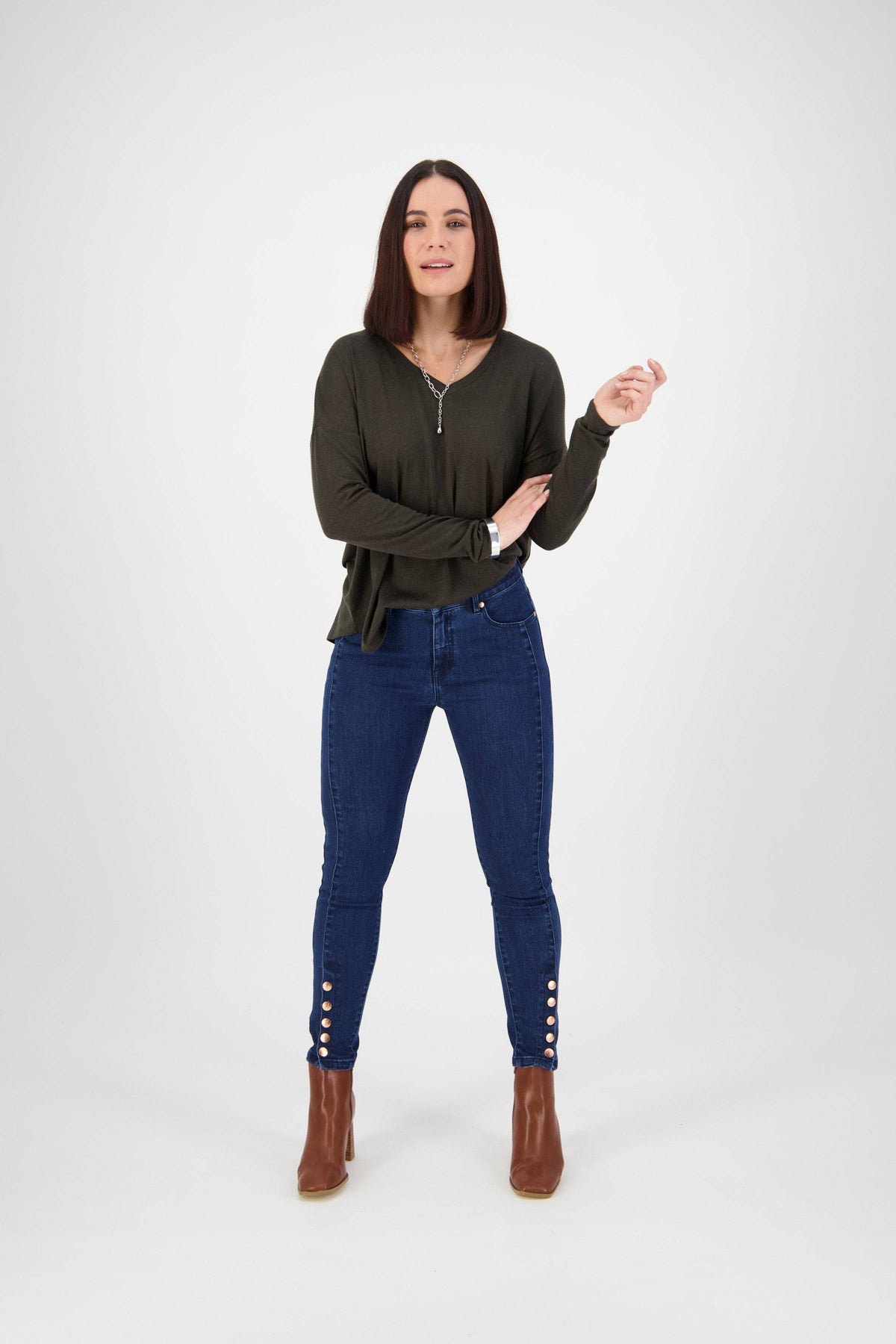 Woman with straight dark hair wears a dark green long-sleeve top, Vassalli Ankle Grazer Jean - Ink Denim, and brown ankle boots, standing against a plain white background.