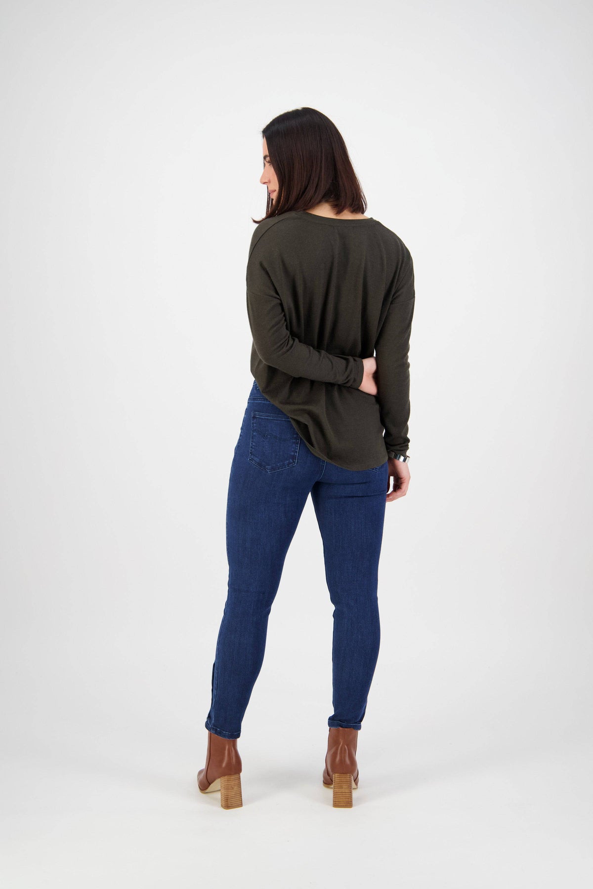 Woman facing away from the camera wears a dark green long-sleeve shirt and Vassalli Ankle Grazer Jean - Ink Denim, paired with brown heeled boots, in a simple white studio setting.