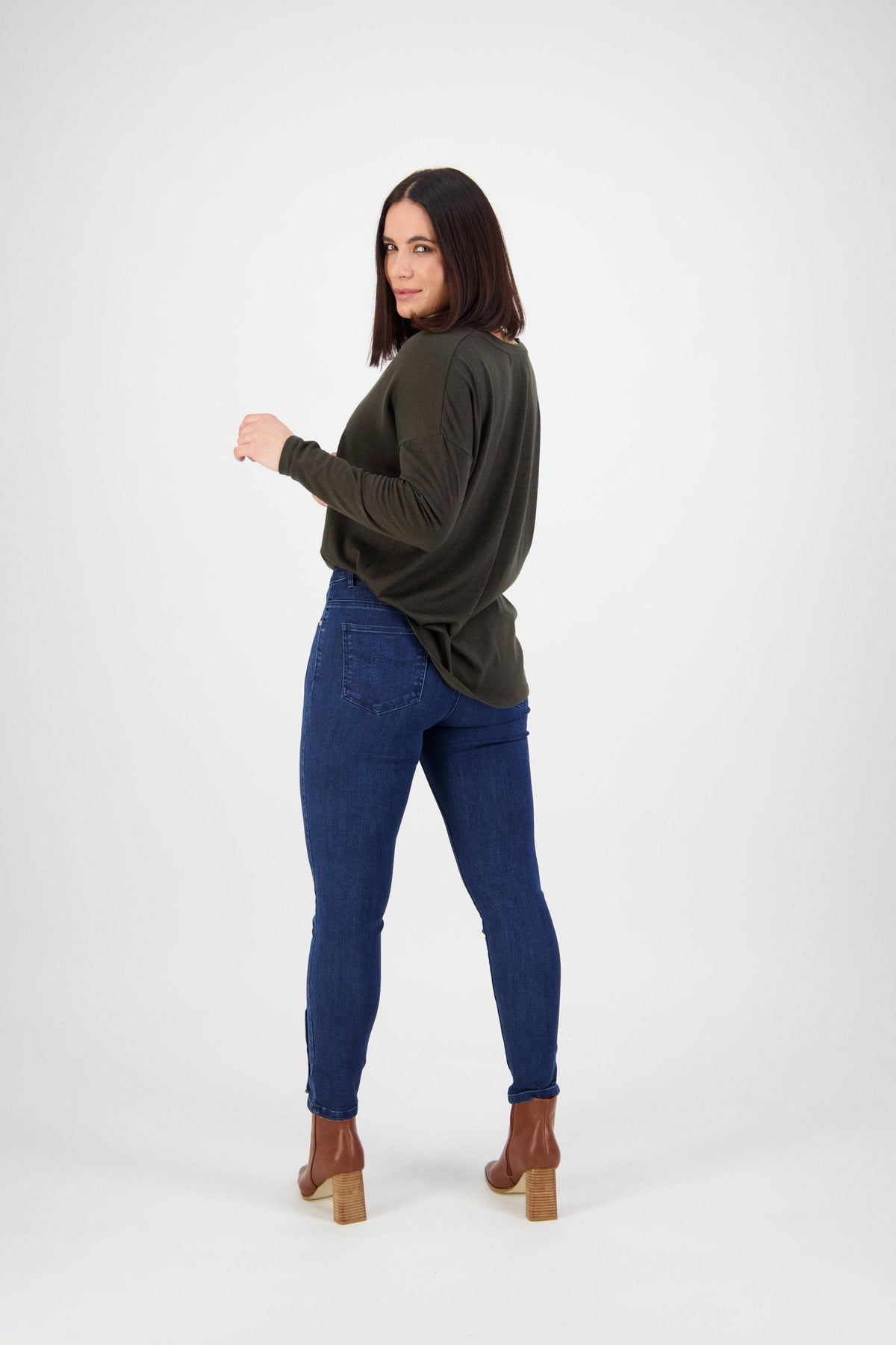 A woman with straight dark hair poses with her back turned, looking over her shoulder. She wears a dark green sweater, Vassalli Ankle Grazer Jean - Ink Denim, and brown heeled boots against a plain white background.