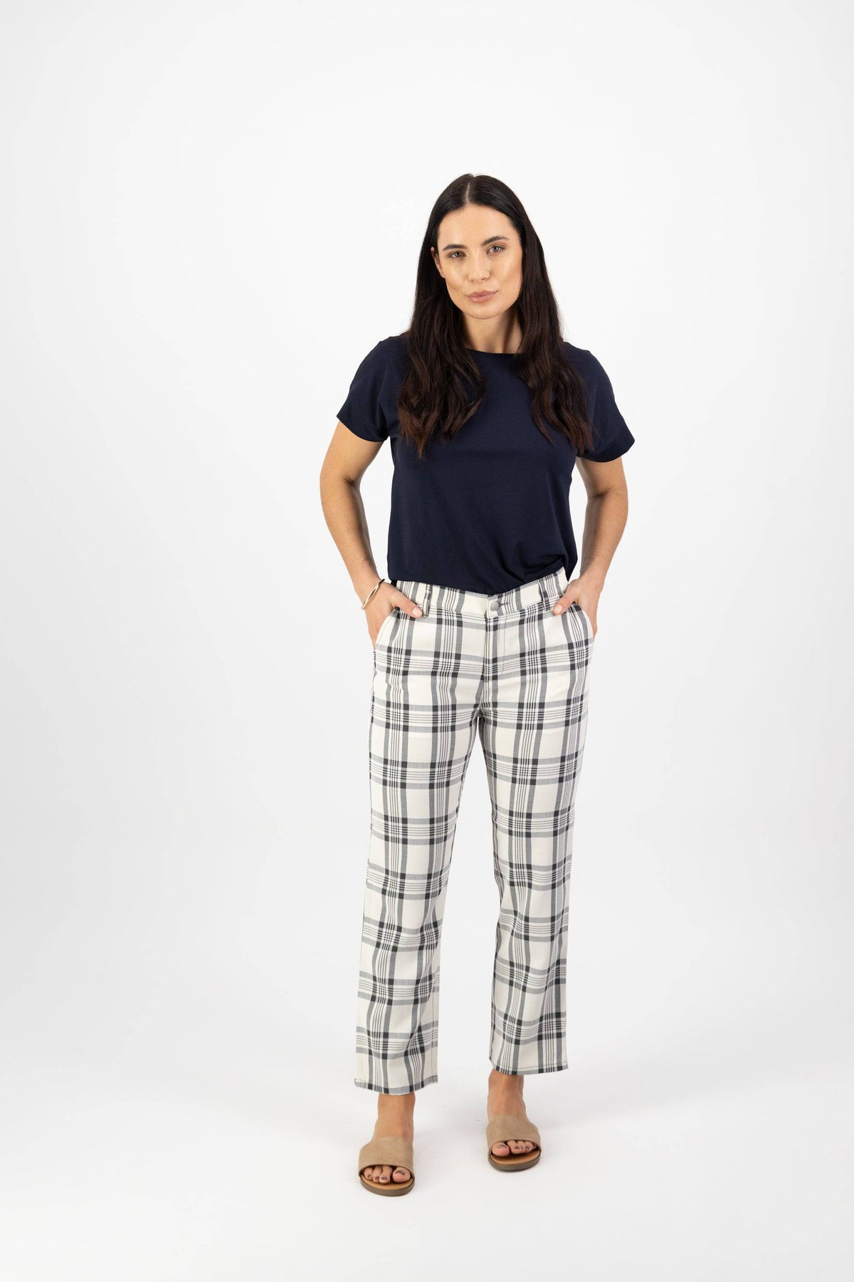 A woman with long dark hair stands against a white background wearing a navy t-shirt, Vassalli 7/8 Pant with Fly in Slate/Off White, and sandals. Her hands are in her pockets.