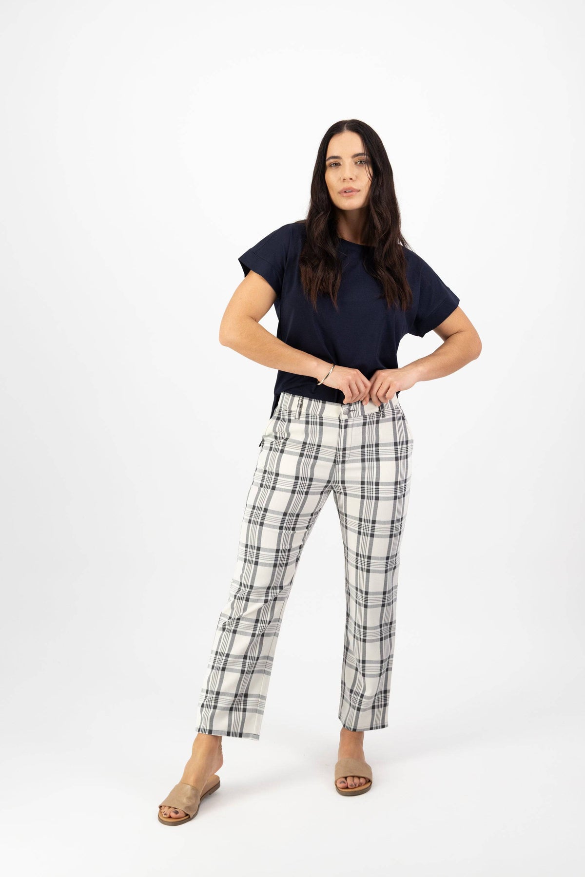 A woman with long dark hair poses against a white background, wearing a navy short-sleeve top and Vassalli 7/8 Pant with Fly in Slate/Off White, styled with tan open-toe sandals. She stands confidently with her hands on her waistband.