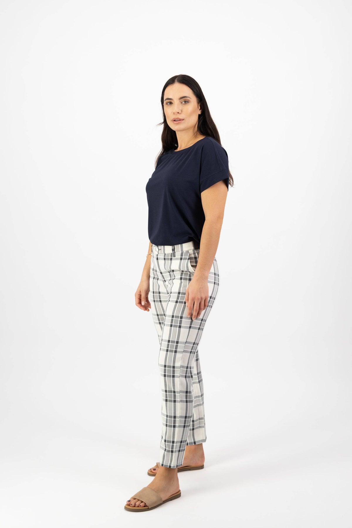 A woman with long dark hair stands confidently against a white background, wearing a navy short-sleeve top and Vassalli 7/8 Pant with Fly in Slate/Off White plaid. The slim pants feature stretch fabric for comfort.