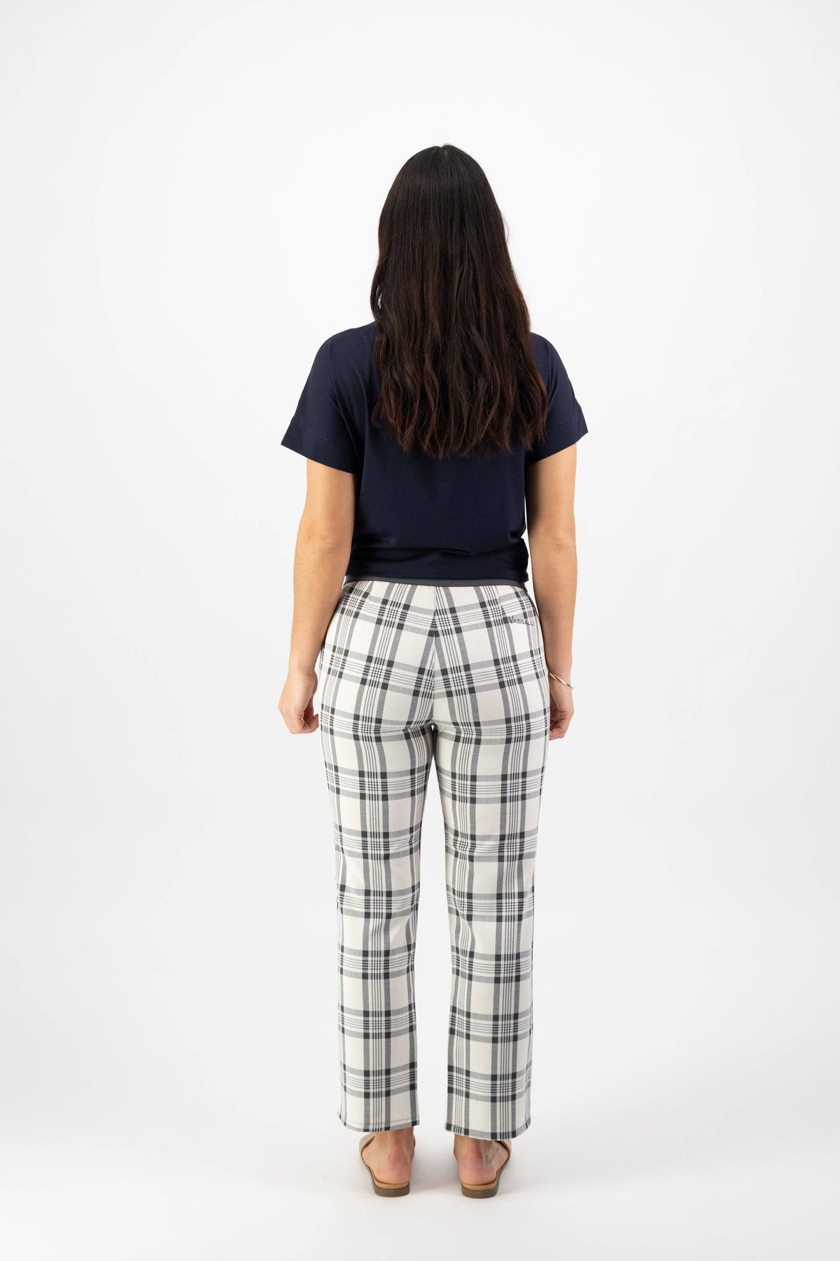 A woman with long brown hair wearing a navy t-shirt and Vassalli 7/8 Pant with Fly in Slate/Off White stands facing away from the camera against a plain white background.