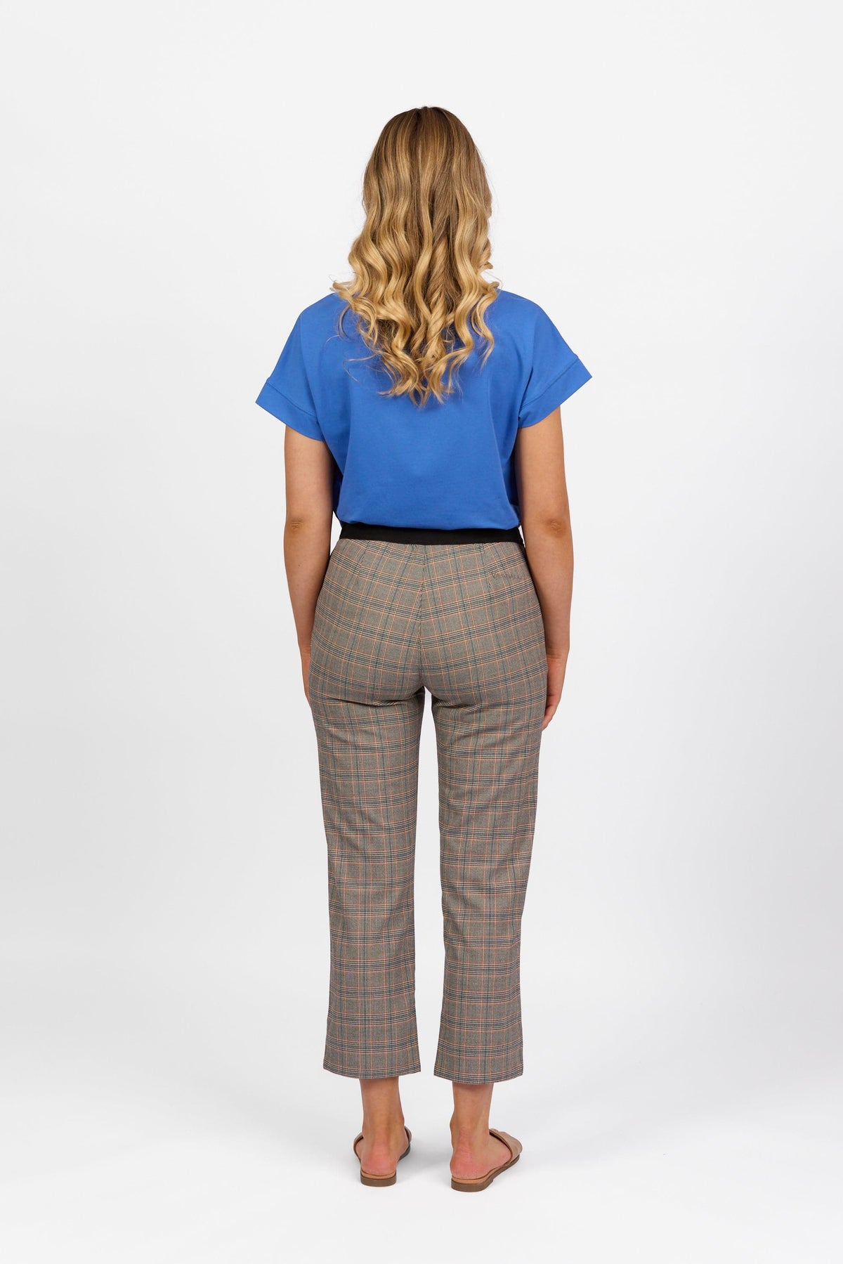 A person with long wavy hair, in a blue short-sleeve top, Vassalli 7/8 Pant with Fly in Tangerine Check, and sandals, stands facing away from the camera against a plain white background.