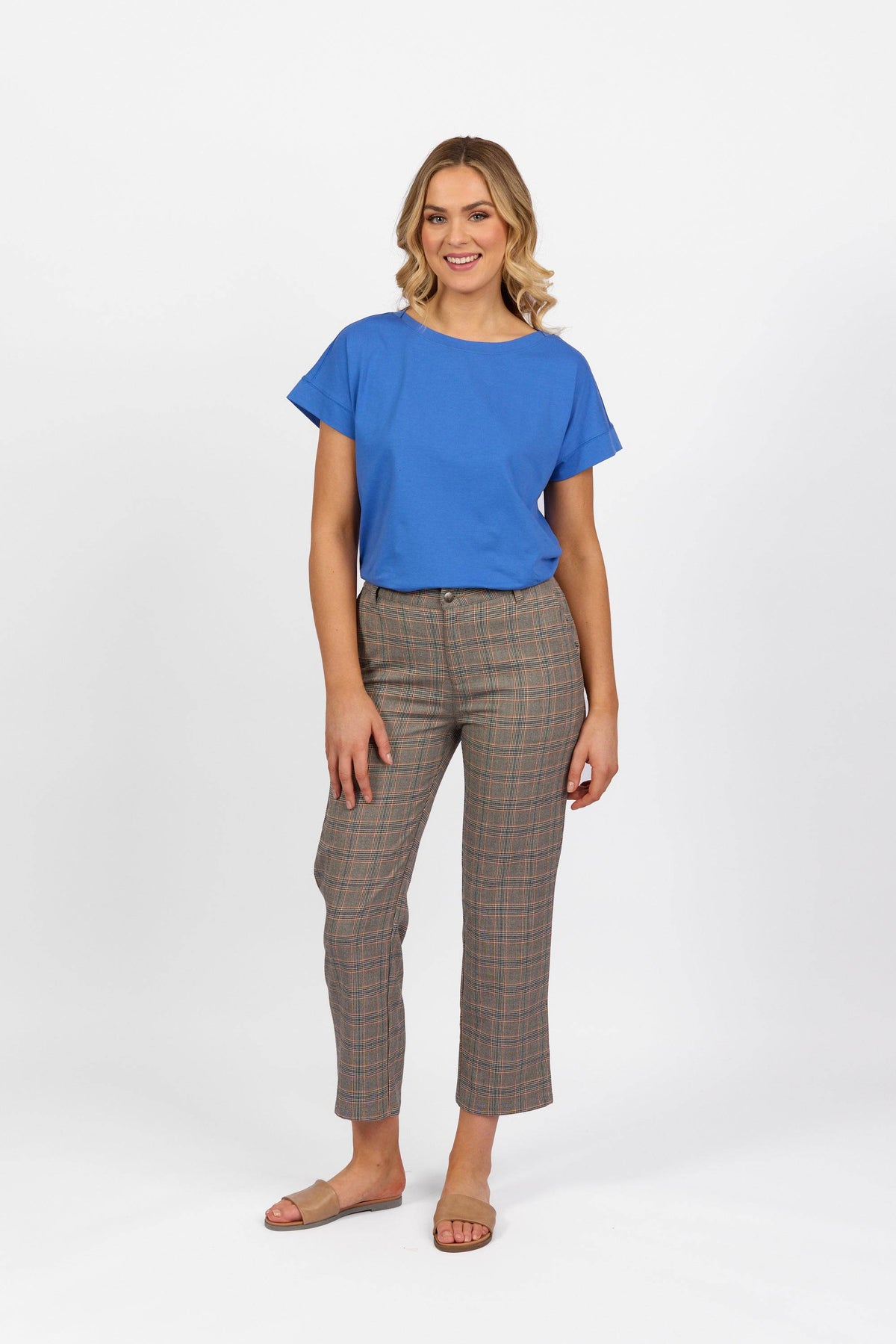 A woman smiles against a white background, wearing a bright blue short-sleeve top, Vassalli 7/8 Pant with Fly in Tangerine Check, and beige slide sandals.