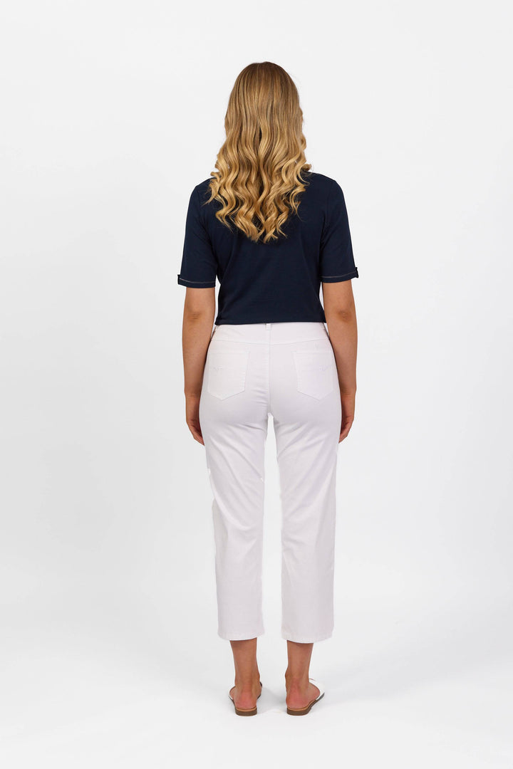 A woman with long wavy blonde hair, seen from behind, wears a navy short-sleeve top, Vassalli Slim Leg 7/8 Pant in white, and flip-flops against a plain white background.