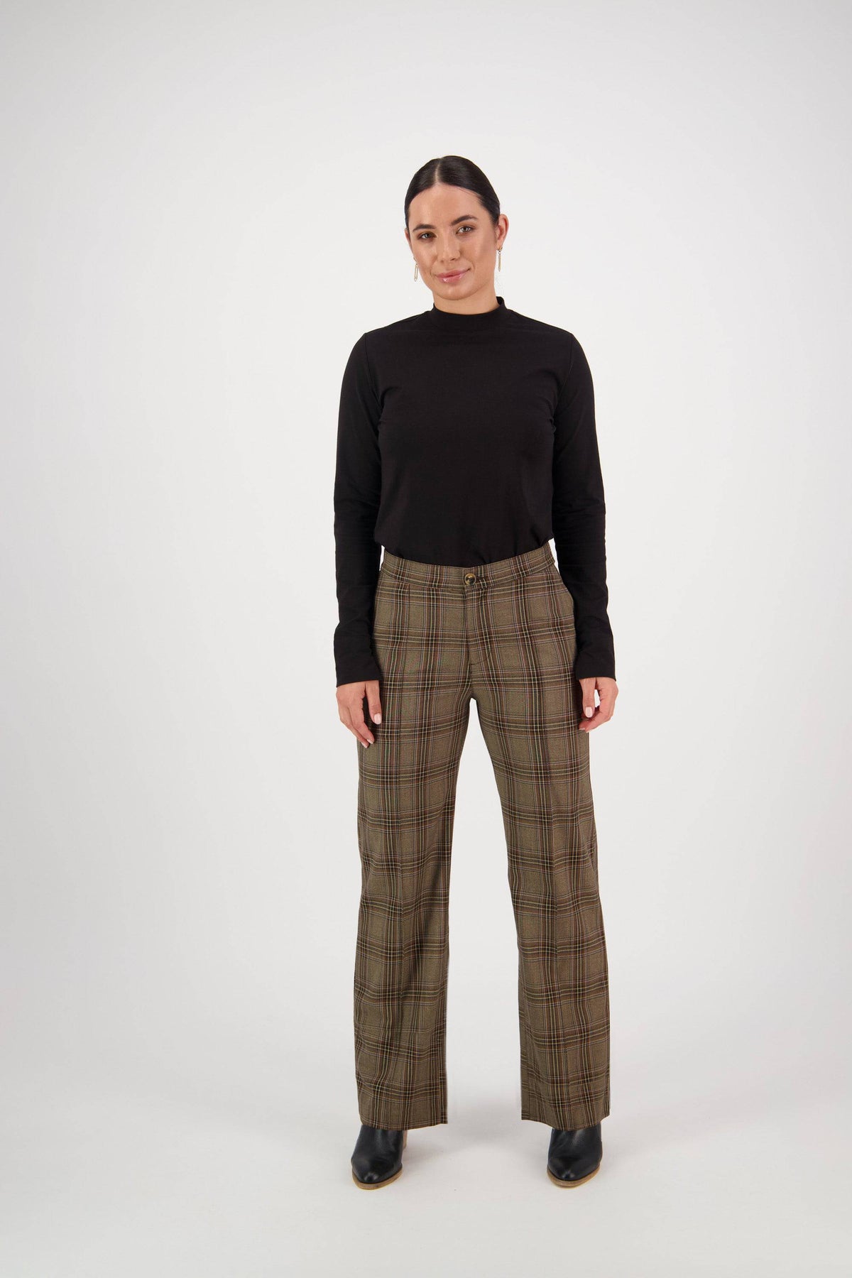 A person faces forward against a white background, wearing a black long-sleeve top and Vassalli Wide Leg Dress Pant - Britain in brown plaid with an elastic back waistband, paired with black shoes.