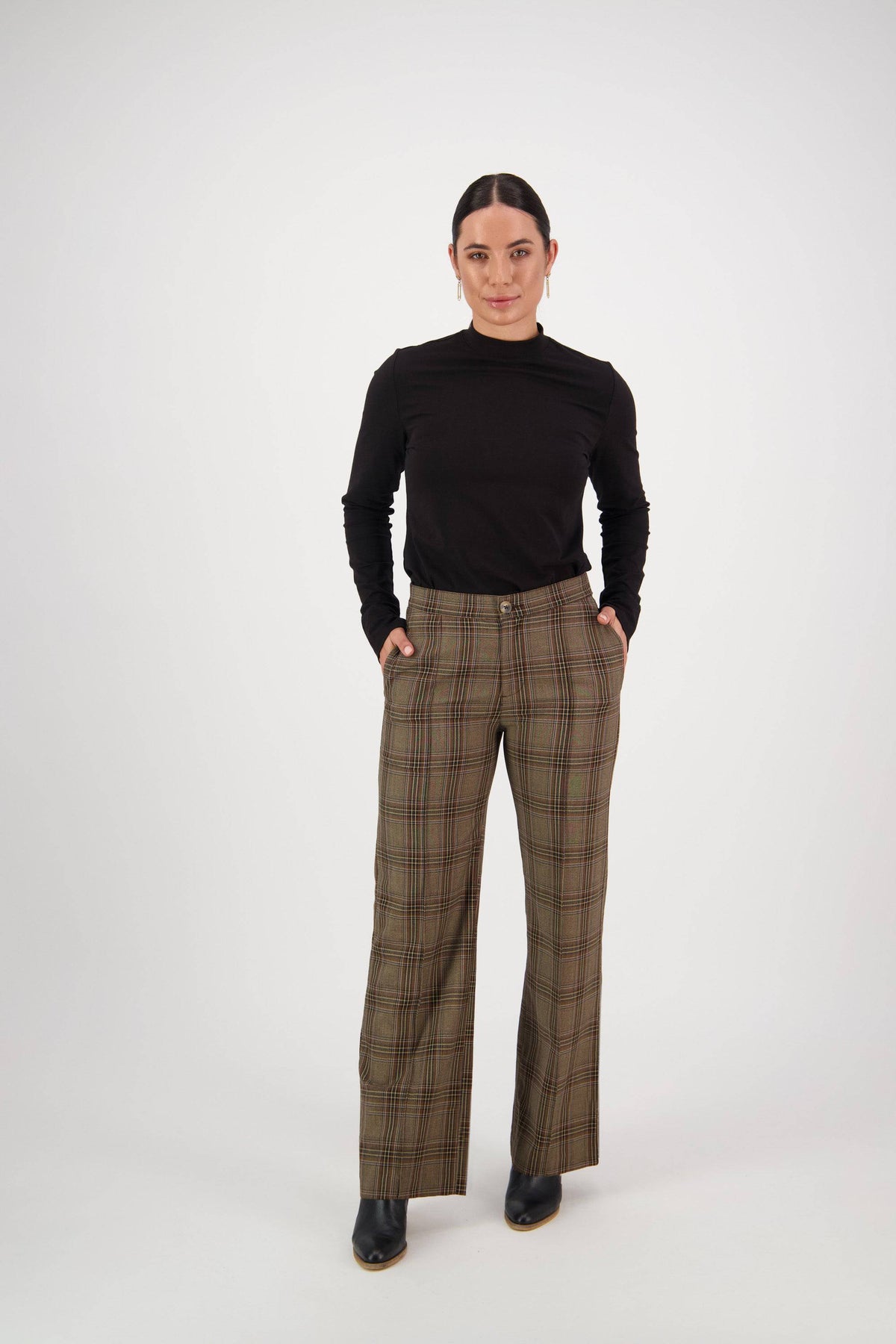 A person stands against a white background, wearing the Vassalli Wide Leg Dress Pant - Britain with an elastic back waistband and a black long-sleeve top, hands in pockets and a neutral expression.