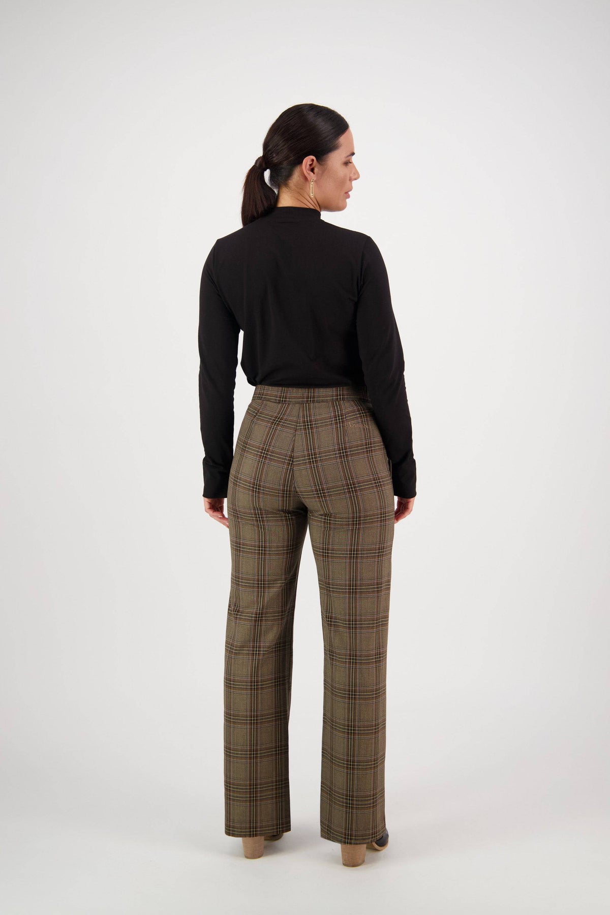 A woman with a low ponytail faces away, wearing the Vassalli Wide Leg Dress Pant - Britain in high-waisted brown plaid and a black long-sleeve top, against a plain white background.