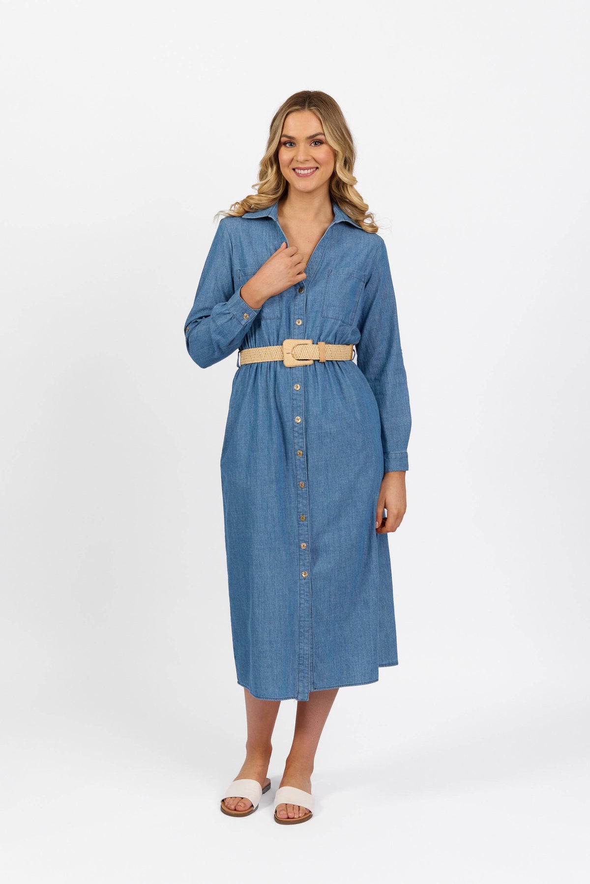 A woman smiles in a Vassalli Long Sleeve Dress - Blue Wash, styled with a beige belt and white slide sandals, standing against a plain white background.